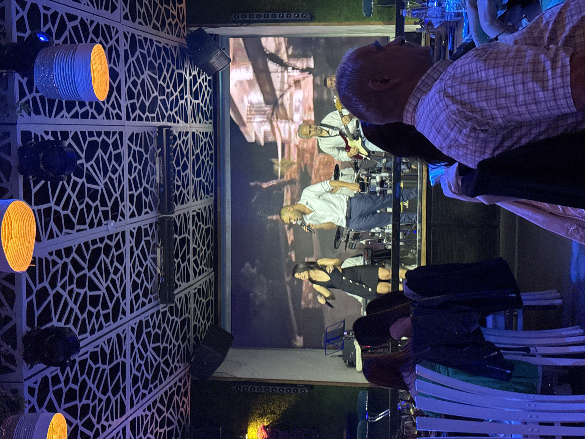 The scene depicts a live music performance, likely a projection on a screen. A man with a microphone is visible, accompanied by a guitarist and a female vocalist. The venue appears to be outdoors with decorative white latticework and colored lighting fixtures on the left. In the foreground, seated patrons watch the performance. There is no identifying information to conclusively confirm this photo was taken in Plovdiv, Bulgaria.