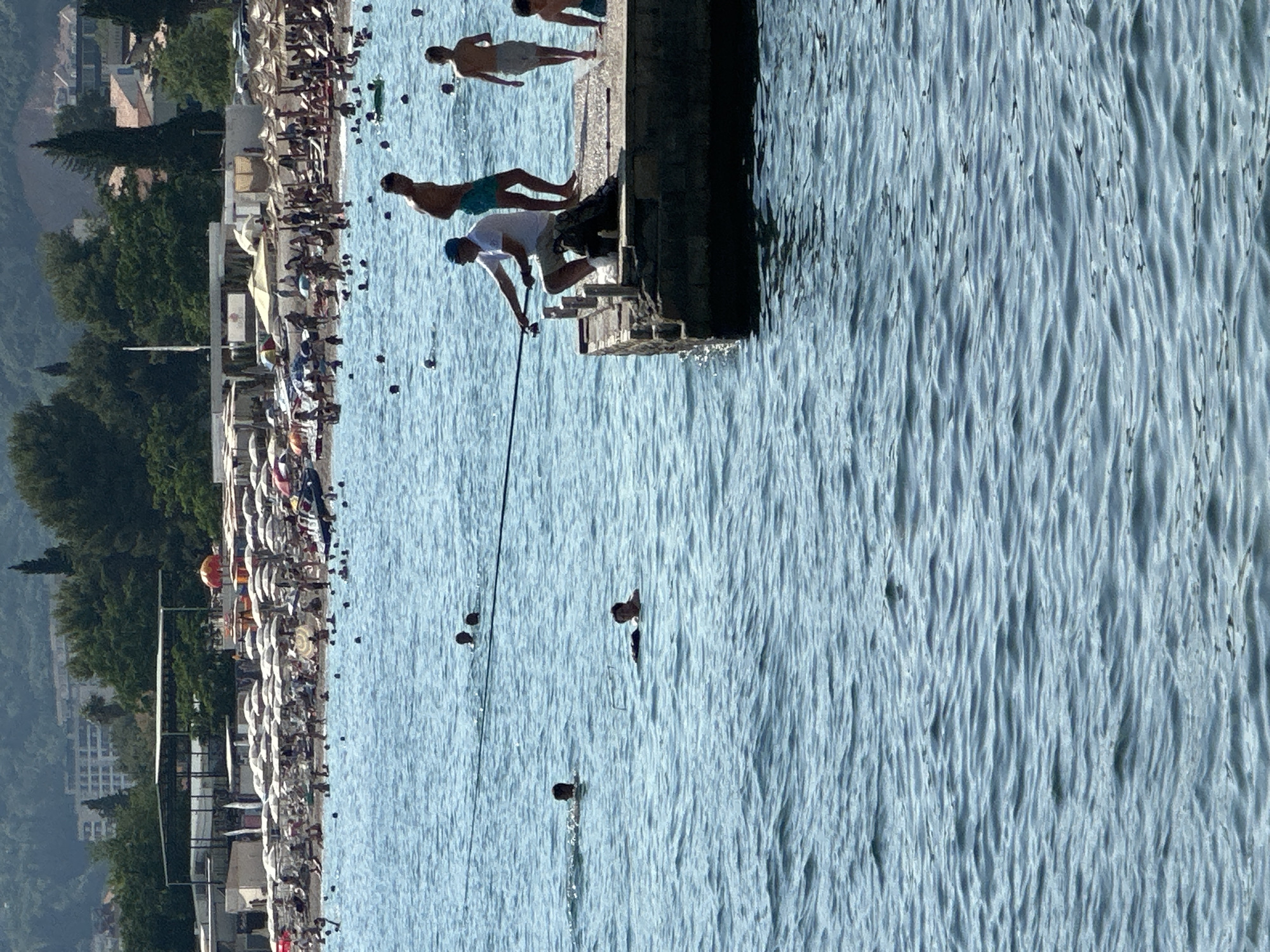 The image depicts a sunny day at a crowded beach in Budva, Montenegro. In the foreground, a man is fishing from a concrete pier. He is wearing a white shirt and beige shorts, and is holding a fishing rod. Behind him, on the pier, are two young men, one with dark hair and wearing blue shorts, and the other with lighter hair and wearing grey shorts. In the water, several people are swimming. The beach is lined with numerous rows of white and beige umbrellas, providing shade for beachgoers who are seated on chairs or lying on towels. Buildings and lush green trees form the backdrop of the scene, with more structures visible on the hills in the distance. The sea is a clear blue with gentle waves.