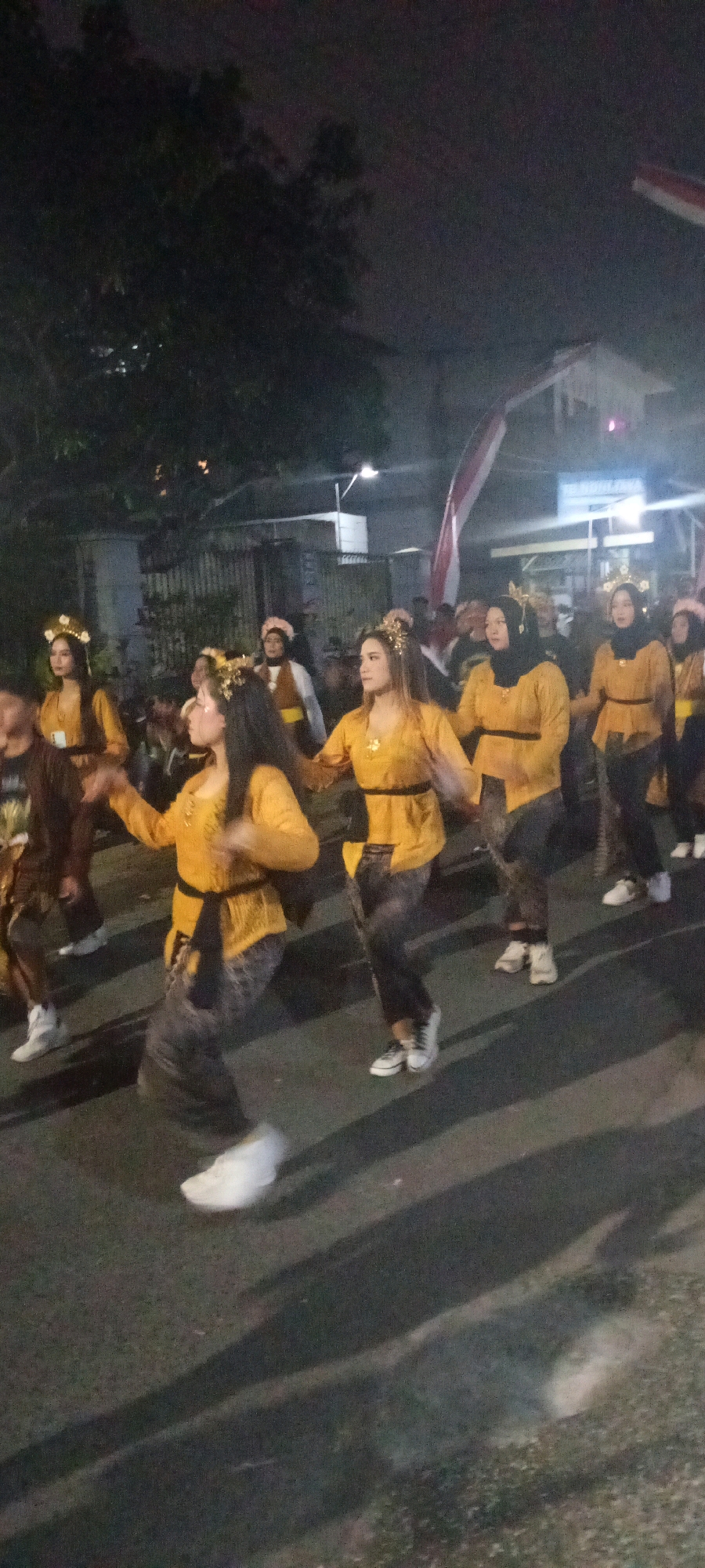 A group of women are parading at night, dressed in traditional yellow outfits with decorative headpieces. They are walking on a paved road, with trees and buildings visible in the background. The image appears to be taken in a city, but the specific location is unknown. The women are the primary subjects of the photograph, and their attire suggests a cultural event or performance. The lighting is dim, indicating it is nighttime, and shadows are cast on the road.