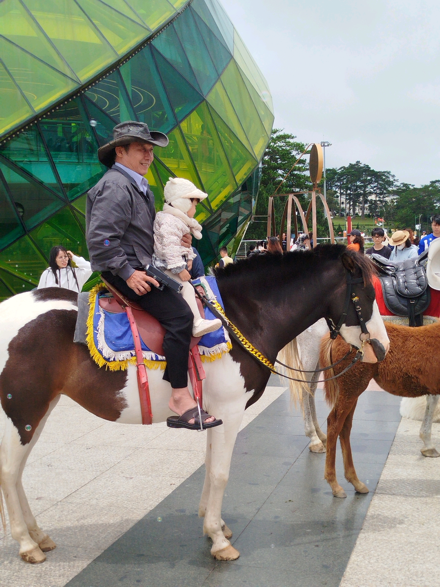 The image depicts a scene in Da Lat, Vietnam, in front of a distinctive, modern building with a faceted, green glass exterior. In the foreground, a man wearing a cowboy hat and a grey jacket is riding a brown and white piebald horse. He is holding a young child, who is dressed in a white outfit and hat, on his lap. The man is holding a black handgun in his right hand. The horse is equipped with a saddle that has colorful blue and yellow trim. To the right of the piebald horse, a smaller, brown horse stands, and further to the right, another horse's hindquarters are visible. Several other people are present in the background, including a woman in a white jacket standing to the left of the horse. The ground is paved with large, grey and white tiles. The overall atmosphere appears to be a tourist attraction, given the presence of horses for rides and the unique architectural landmark.