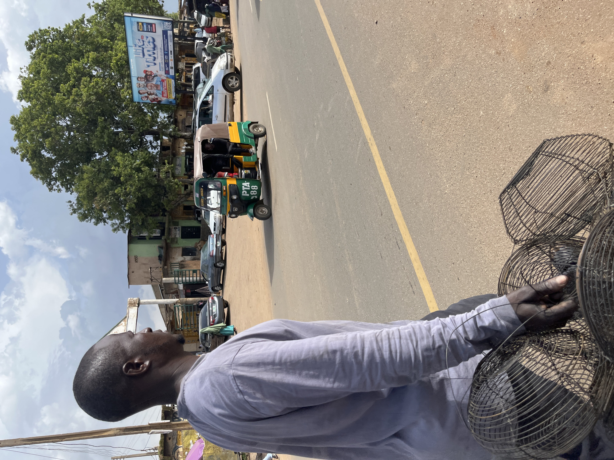 The image depicts a street scene in Jos, Nigeria, under a partly cloudy sky. In the foreground, a man wearing a light purple long-sleeved shirt is seen from the waist up, facing to the right, holding several wire baskets. His skin is dark, and he has short, dark hair and a beard. His right hand is visible, touching one of the baskets.
Further in the background, a green and yellow auto-rickshaw, identified by the license plate "PTA 88", is positioned on the asphalt road. A person is seated in the driver's seat. Several other vehicles, including a silver minivan and a dark sedan, are also present on the road.
Buildings line the street, with one appearing to be a shop or small establishment with a light green facade. A large billboard displaying "Life Times" and images of several men is visible in the middle ground, behind the auto-rickshaw and trees. People are scattered throughout the scene, some near the vehicles and buildings. The road is paved with asphalt and marked with a yellow line on the right side. The environment suggests an urban or semi-urban setting within Jos.