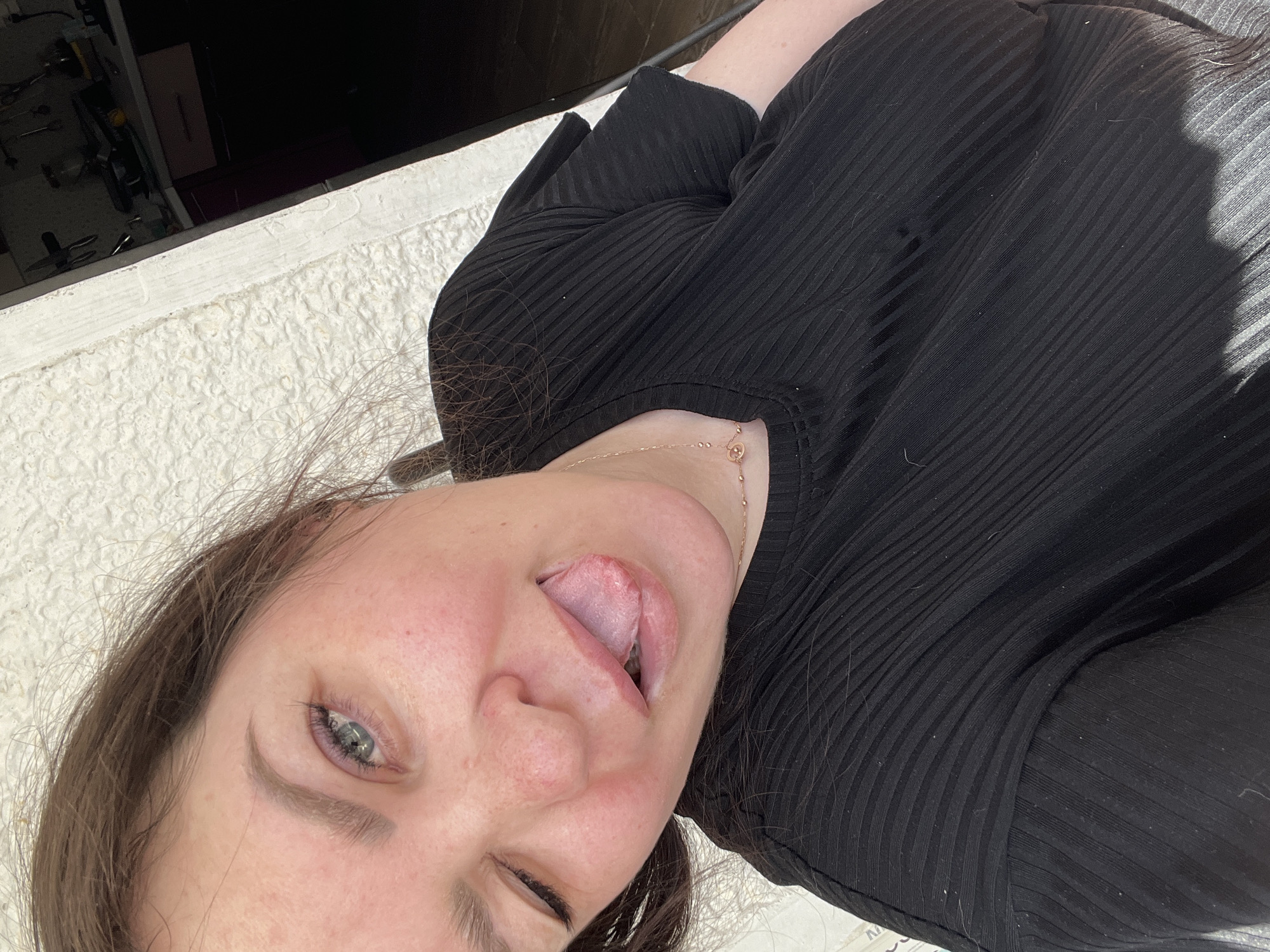 A woman with light skin and brown hair is sticking her tongue out and winking. She is wearing a black, ribbed shirt and a gold necklace. The background is a blurry mix of white concrete and a dark interior. The location of Noviy, Russia is not discernible from the image.
