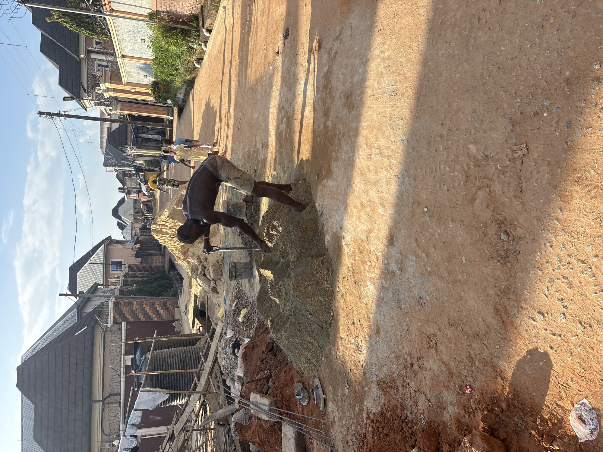 A man is working with a shovel in a pile of sand on a dirt road in Obosi, Nigeria. He is shirtless and wearing shorts. In the background, there are several houses with tiled roofs and a few children walking down the road. The sun is shining, casting long shadows. Construction materials, including rebar and a bag of cement, are visible near the man. There are also water tanks and scaffolding on the left side of the image.