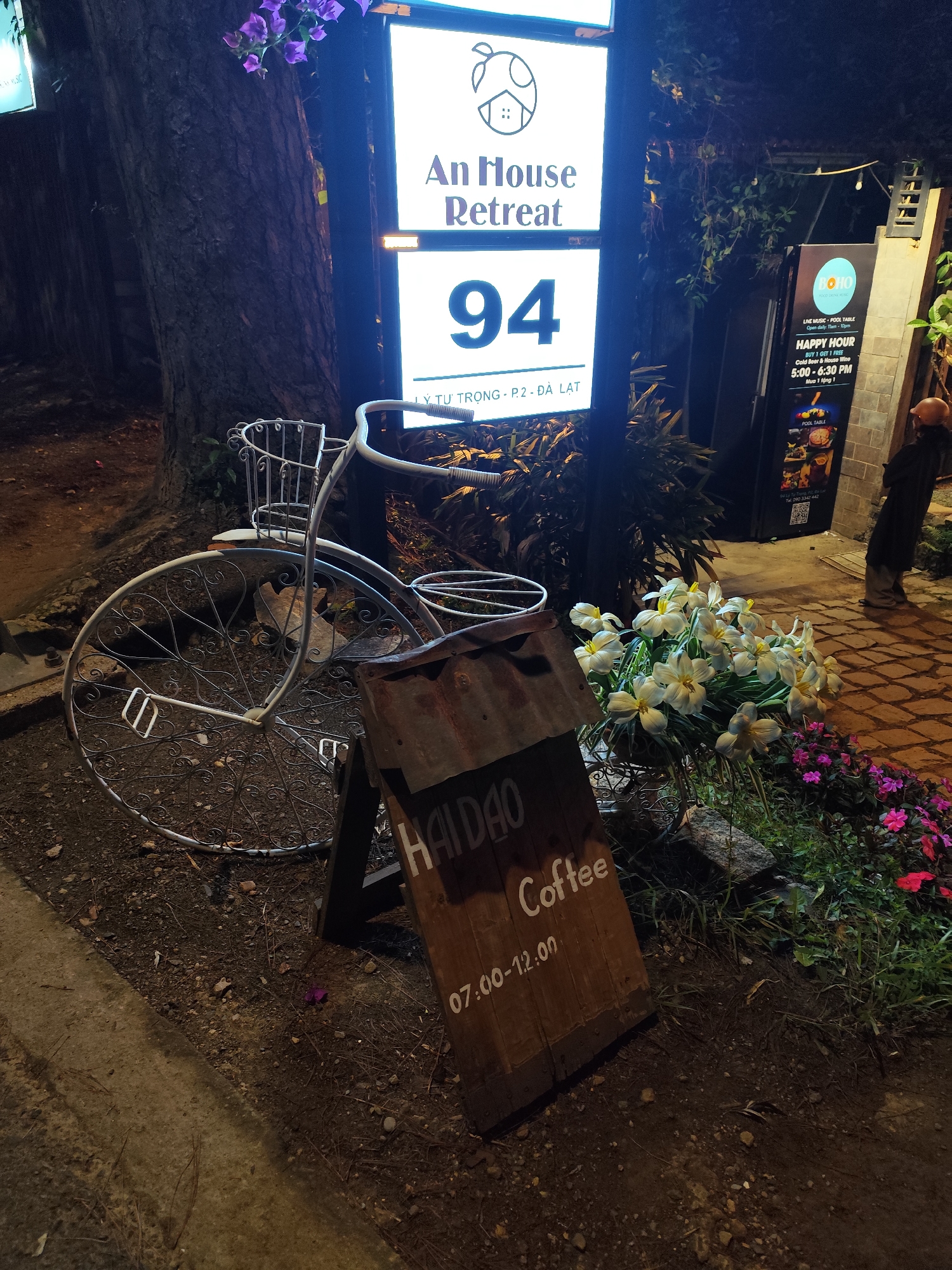 The image shows a nighttime scene outside "An House Retreat" at number 94 Ly Tu Trong street, Ward 2, Da Lat, Vietnam. A decorative, vintage-style bicycle stands next to a wooden sign reading "Hai Dao Coffee" with operating hours 07:00-12:00. Flowers are planted next to the bicycle and sign. In the background, a sign for "Boho" advertises live music, pool table, and a happy hour from 5:00-6:30 PM. A person wearing a dark outfit is standing by the Boho sign. A large tree trunk is visible to the left of the retreat's sign.