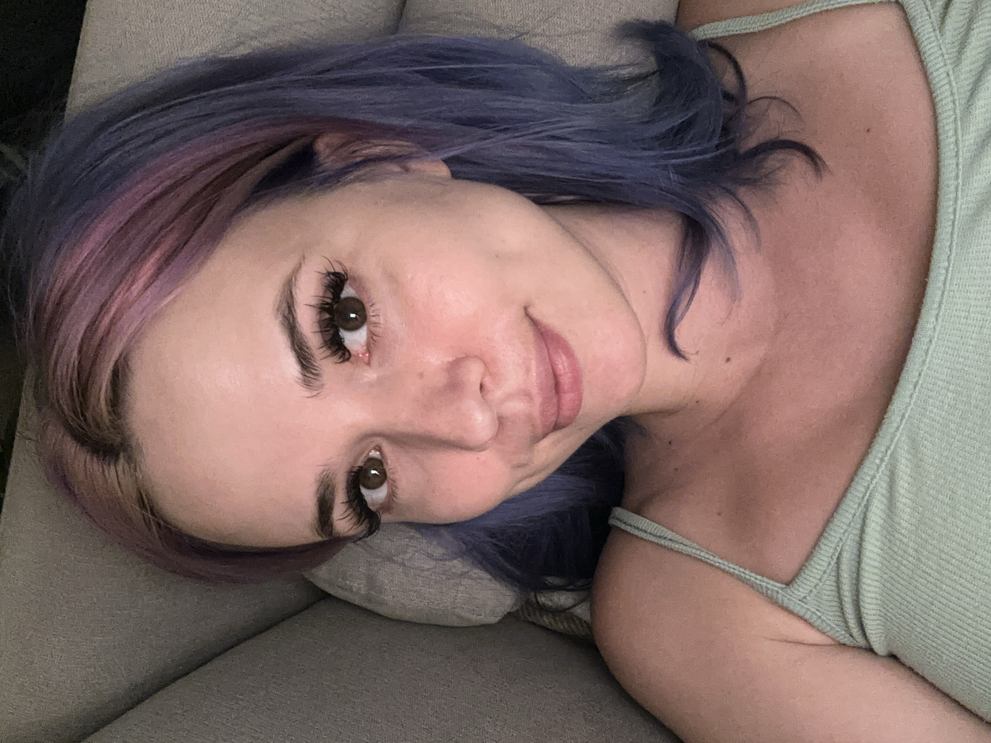 A woman lies on a grey upholstered surface, possibly a sofa or bed. She has fair skin, brown eyes, and multicolored hair, featuring hues of pink and blue-purple. The woman is wearing a light green tank top with thin straps. The photograph is taken from an overhead perspective, showing her face turned towards the camera.
There is no information in the image that could place it in Gijón/Xixón, Spain.