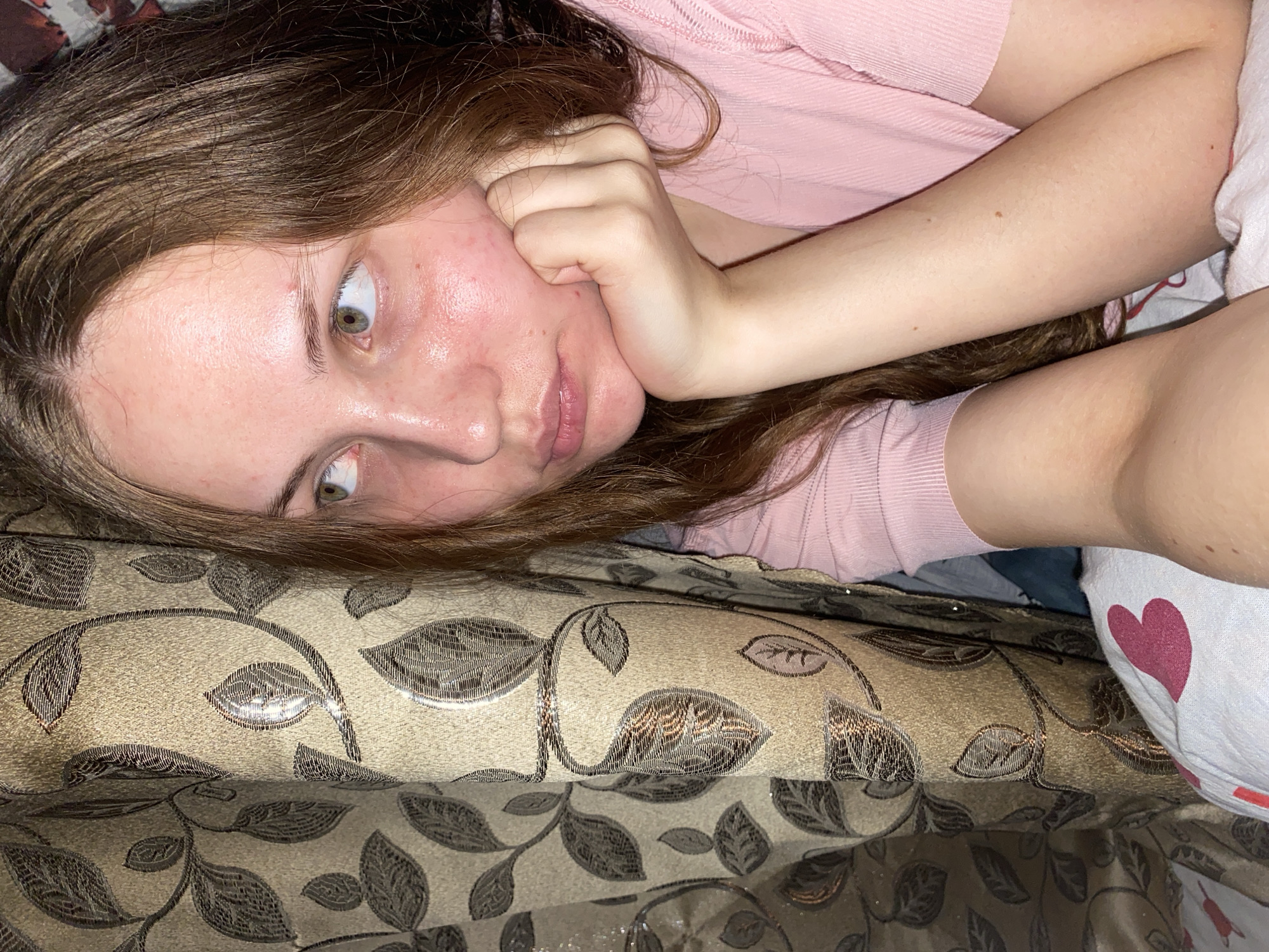 The image shows a young woman with long brown hair, resting her head on her hand. She is wearing a pink shirt and appears to be looking off to the side. The background features a patterned curtain and bedding with a heart design. The overall scene is intimate and appears to be indoors, possibly in a bedroom. There is no visible indication of the location being in Вассята, Russia.