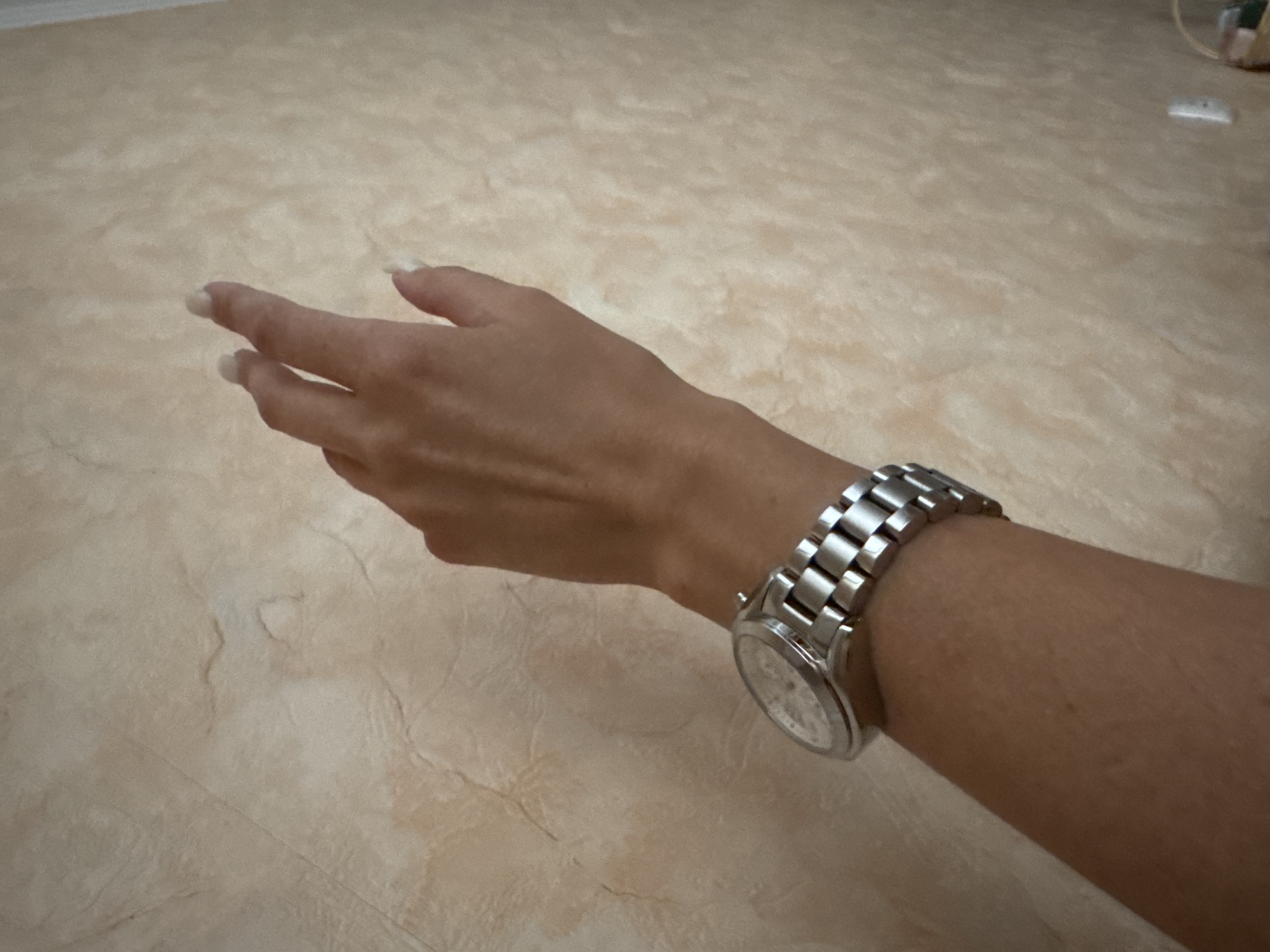 A person's arm, adorned with a silver wristwatch, is extended towards a textured, pale peach-colored wall. The hand is positioned with fingers slightly curled, pointing towards the upper right portion of the frame. The wall exhibits an irregular, mottled pattern with subtle variations in shading. No specific location in Uman, Ukraine is discernible from the image.