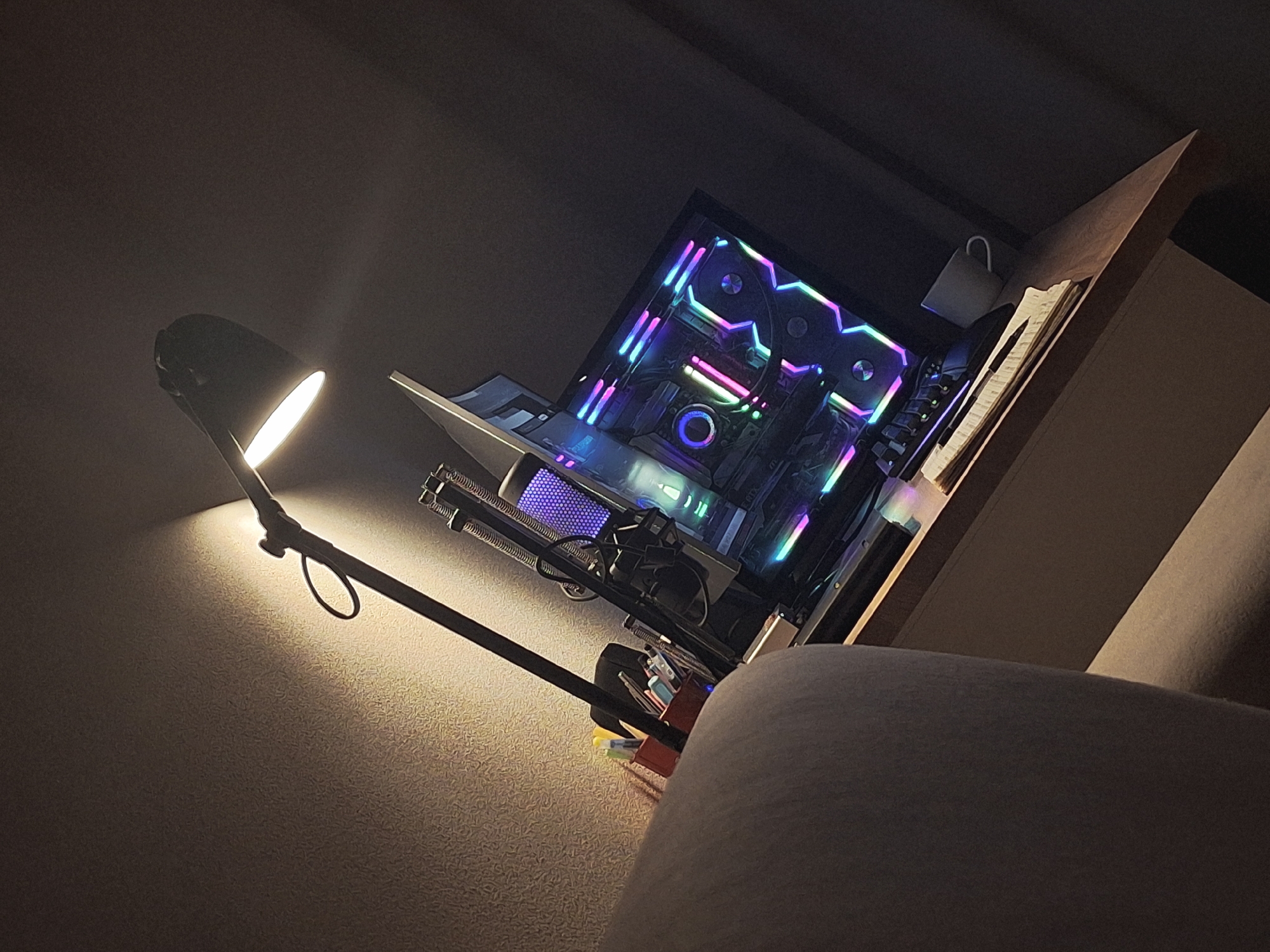 The image presents a desk setup in a dimly lit room. A desk lamp illuminates the wall to the left, casting a bright spotlight. The focal point is a computer tower with glowing RGB lights, showcasing internal components. The tower sits on the desk alongside a desk organizer filled with pens and other stationery. A mug hangs from the shelf above the desk next to a notebook. A portion of the chair is visible in the foreground. The image lacks any information about Chaikovsky, Russia.