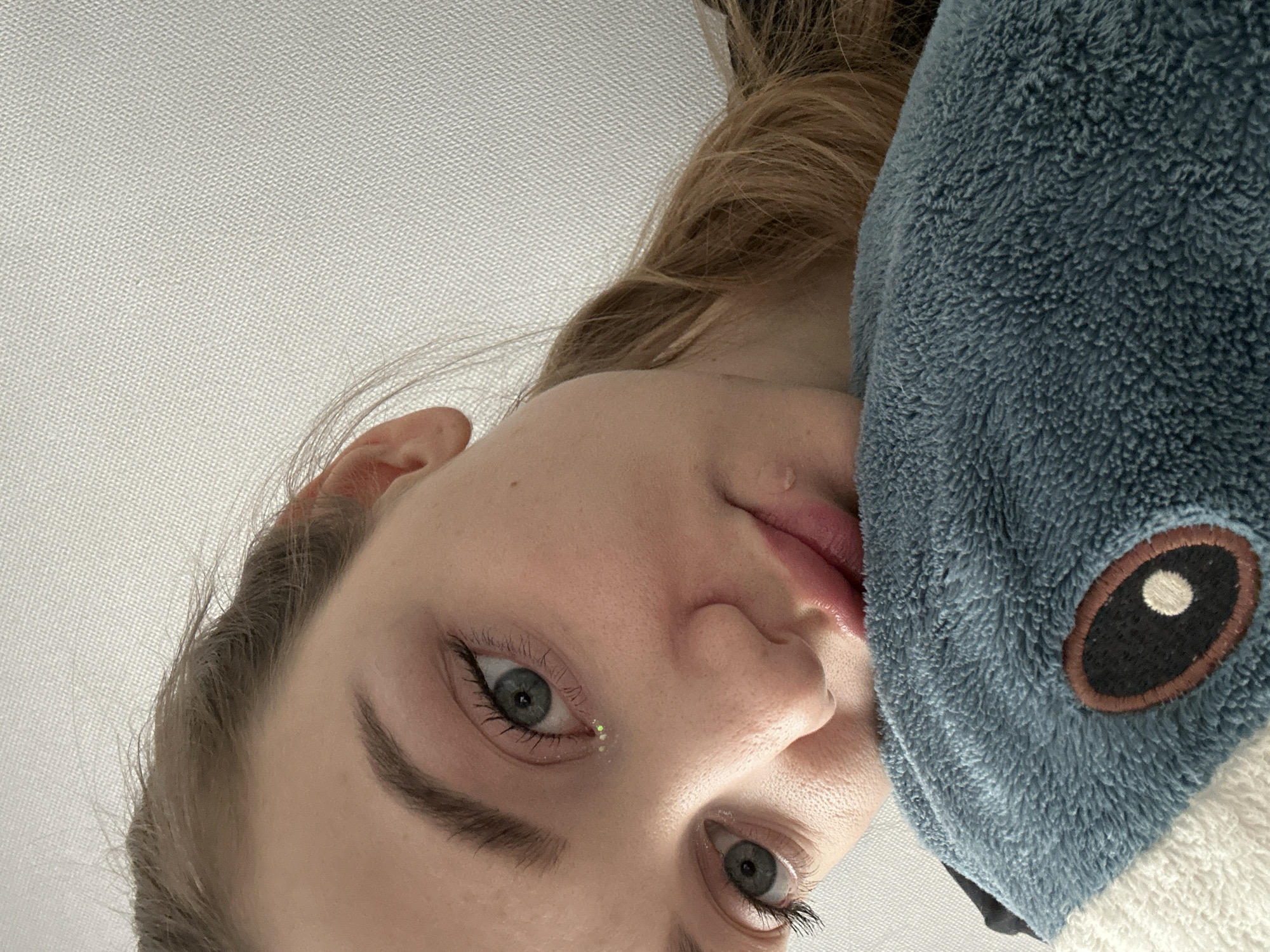The image is a close-up, high-angle portrait of a young person, rotated 90 degrees clockwise, giving the impression of looking up at them. The subject has fair skin, light brown hair, and blue/grey eyes with long eyelashes. A small, iridescent sparkle or glitter is visible near the inner corner of their left eye. Their lips are slightly parted.
On the right side of the frame, the person is wearing a fluffy, deep blue garment, likely a hooded blanket or a kigurumi. An embroidered eye, consisting of a dark brown oval with a white circular highlight, is visible on this blue material, suggesting a cartoon or animal character. The background behind the person is a light, off-white textured surface, possibly a ceiling or fabric.
The overall scene is intimate and casual. The person's expression appears neutral or slightly contemplative. The lighting is soft and even, suggesting an indoor setting with good ambient light. There are no explicit location cues within the image itself. No text is visible. The provided location context for this media file is Kazan, Russia.