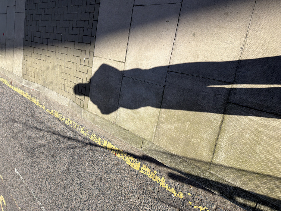 The image depicts an outdoor urban environment, specifically a street and pavement under strong sunlight, casting long shadows. The scene is consistent with a location in London, United Kingdom, identifiable by the continuous yellow line road marking adjacent to the curb.
The foreground features a pavement composed of light-toned, rectangular paving slabs, some exhibiting a textured, possibly basketweave, pattern. To the left of the pavement, a dark grey asphalt road surface with visible aggregate is present. A concrete curb separates the pavement from the road. On the road, a continuous yellow line runs parallel to the curb, indicating a parking restriction. Further into the road, a faded, broken white line is discernible.
Two primary shadows are prominent: an elongated shadow of a human figure is cast diagonally across the pavement, originating from the lower right and extending towards the center. The shadow's posture suggests the individual is standing upright, potentially holding an object at chest level or having their arms folded. A second distinct shadow, displaying a branching pattern characteristic of a bare tree or tree branch, is cast across the asphalt road and partially onto the curb. Additionally, a broad, dark shadow, likely from an adjacent building or structure, partially covers the pavement on the right side of the frame. No actual people or specific objects are visible, only their cast shadows. The lighting indicates direct sunlight from a relatively low angle.