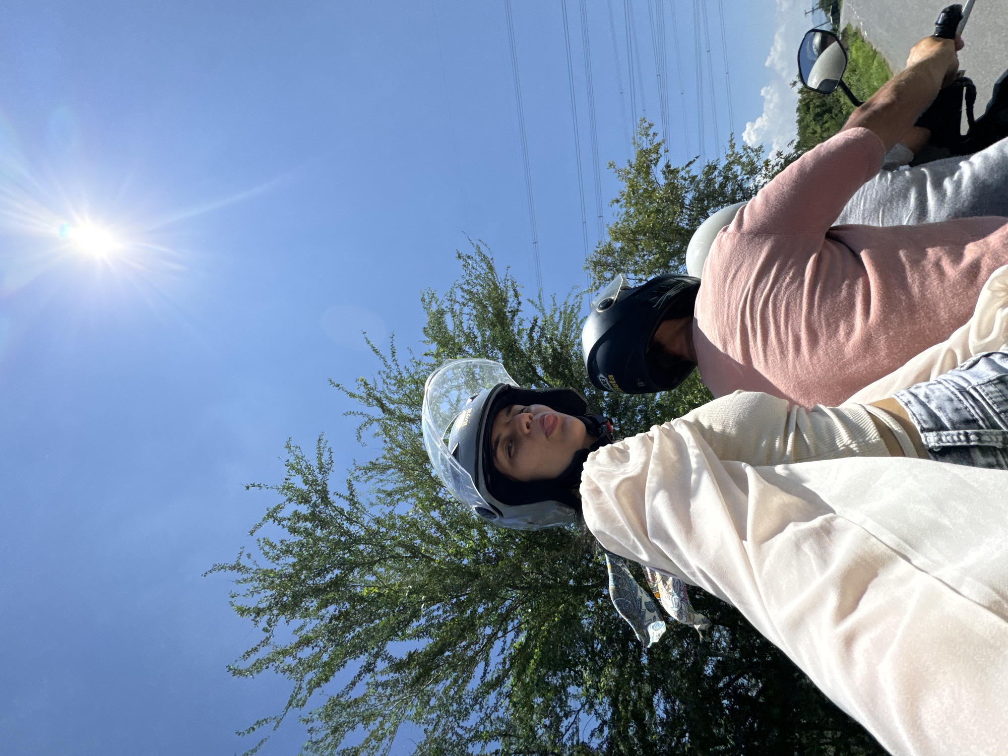 The image depicts two individuals riding on a motorcycle under a bright, clear blue sky with the sun high and shining intensely. The person in the foreground, on the left, is a woman wearing a black helmet and a light-colored, sheer long-sleeved top over a ribbed tank top, and distressed jeans. She is looking towards the camera and sticking her tongue out. A patterned scarf is visible around her neck. Behind her, another person, likely male, is wearing a black helmet and a pink t-shirt. A third person, only partially visible, is wearing a gray t-shirt and a white helmet. The background consists of lush green trees, power lines stretched across poles, and a paved road curving to the right. The overall setting suggests a rural or suburban area, potentially on the outskirts of Hong Son, Vietnam, given the vegetation and the presence of power lines. The perspective is from the motorcycle, looking forward and slightly to the side.