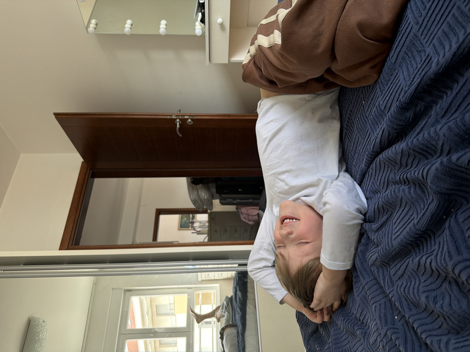 The image displays a young individual, possibly male, with light brown hair, lying supine on a bed in a bedroom in Budva, Montenegro. The person is wearing a white long-sleeved shirt and brown sweatpants featuring light-colored horizontal stripes on the outer thighs. Their arms are bent, with hands clasped behind the head. The individual's eyes are closed, and their mouth is open in a wide expression consistent with laughter or a broad smile.
The bed is covered by a dark blue blanket with a raised, geometric texture. To the left of the bed, a mirrored sliding wardrobe reflects a window with white frames, revealing an exterior building with light-colored walls. The reflection also shows a portion of another bed with a person's bare leg, bent at the knee, visible. To the right of the individual, a dark wooden door stands ajar, opening into an adjacent room. Visible through the doorway are a grey chest of drawers, stacked luggage, and a partially discernible framed picture on the wall. Further to the right in the main bedroom, a white dressing table with a rectangular mirror illuminated by a vertical array of three spherical light bulbs on each side is present. The walls of the bedroom are painted a light, neutral color.
