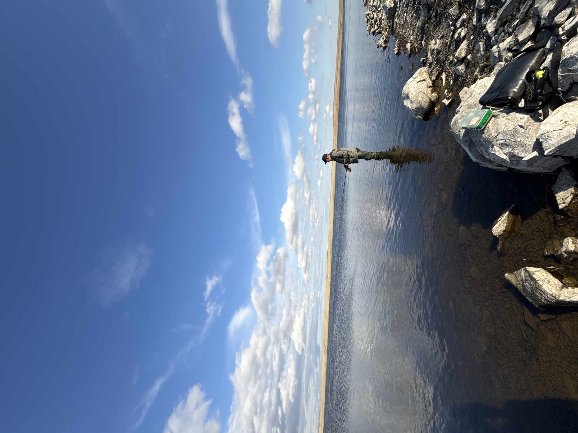 A man is fishing in a lake under a blue sky with scattered clouds. He is wearing a cap, a jacket, and waders. The lake is calm, reflecting the sky and the surrounding landscape, which appears to be a grassy plain. There are rocks on the shore of the lake, and a fishing tackle box and a bag are placed on a rock. The scene is set in Matraevo, Russia.