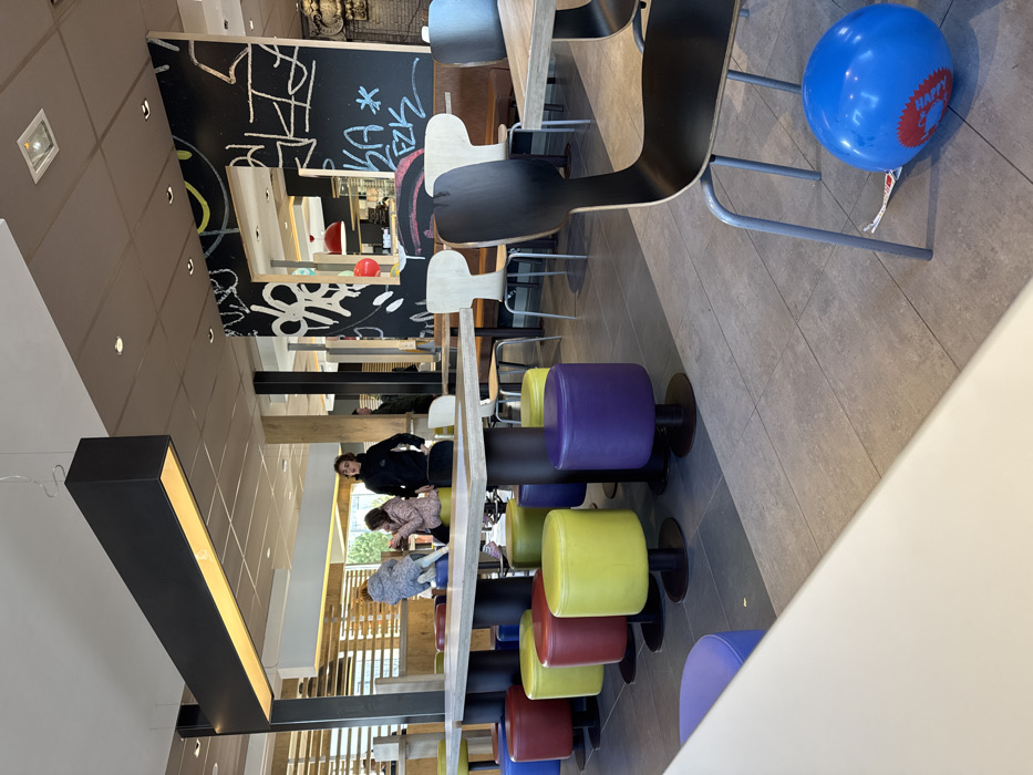 This image depicts the interior of a fast-food restaurant, identified as a McDonald's, located in Oberhausbergen, France. The scene is observed from a slightly elevated perspective, looking towards the mid-ground.
**Environment:**
The dining area features a light grey tiled floor. The ceiling is paneled in a light brown or beige hue, with recessed lighting fixtures. A prominent, elongated black rectangular light fixture with an illuminated underside hangs from the ceiling in the mid-left. Dark pillars are visible, some adorned with white graffiti-style markings and text, including a section with a mirror-like cutout. Blinds or slatted structures are visible through a large window in the far background on the left. Red hanging dome lights are visible in the distant background.
**Seating and Tables:**
In the foreground, the edge of a white table is visible on the left. In the mid-ground, a long, light-colored rectangular communal table with a dark base is surrounded by a row of colorful, round upholstered stools in blue, red, yellow, and purple, each with a dark circular base. Beyond this, various other tables and chairs are arranged throughout the dining area, including light wood-toned chairs with dark backs and brown upholstered booth seating. In the immediate right foreground, the back and metal legs of a dark chair are prominent.
**People:**
Three individuals are situated around the communal table in the mid-ground. On the far left, a young child with light-colored hair, wearing a patterned jacket, is kneeling on a blue stool. Next to the child, a person with light brown hair, wearing a light-colored top, is seated on a yellow stool, looking down at a device in their hands. To their right, a person with dark, curly hair, wearing a black hooded top, stands beside the table, facing towards the left. A black bag rests on the floor next to this individual. Partially obscured figures are visible further in the background.
**Objects and Actions:**
The child on the stool is positioned as if interacting with the table or the person next to them. The seated person is engaged with a personal electronic device. The standing person appears to be observing the area or waiting. On the floor in the bottom right corner, next to the chair, lies a deflated or partially deflated blue balloon with red text, partially visible as "Happy," indicating a "Happy Meal" balloon. A white ribbon is attached to the balloon.