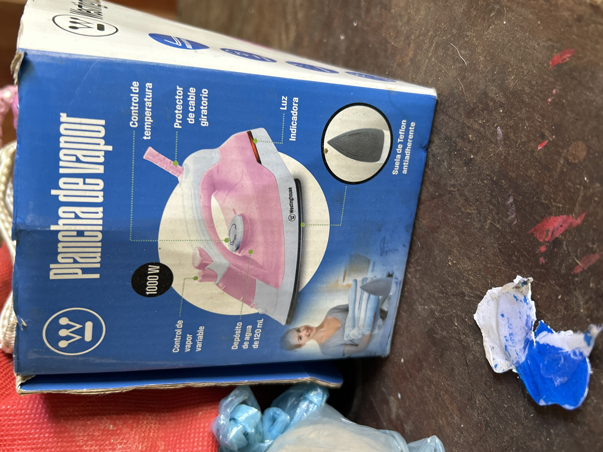 The image shows a close-up of a Westinghouse steam iron box placed on a dark, rustic wooden surface. The box is predominantly blue with white text and graphics. Prominently displayed is the Westinghouse logo, followed by the text "Plancha de vapor" (Steam iron). Various features of the iron are highlighted with dashed lines and Spanish labels: "Control de vapor variable" (Variable steam control), "1000 W" indicating its power, "Control de temperatura" (Temperature control), "Protector de cable giratorio" (Swivel cord protector), "Depósito de agua de 120 mL" (120 mL water tank), "Luz Indicadora" (Indicator light), and "Suela de Teflon antiadherente" (Non-stick Teflon soleplate). A graphic of a pink steam iron is shown in the center, and a smaller circular inset displays a close-up of the iron's soleplate. A woman in a blue shirt is depicted holding folded clothes. The background is out of focus, but hints of red fabric and blue plastic packaging are visible. There is also a piece of blue and white debris on the wooden surface in the foreground. The lighting suggests an indoor setting. There is no information about the location in Santa Ana, Venezuela.