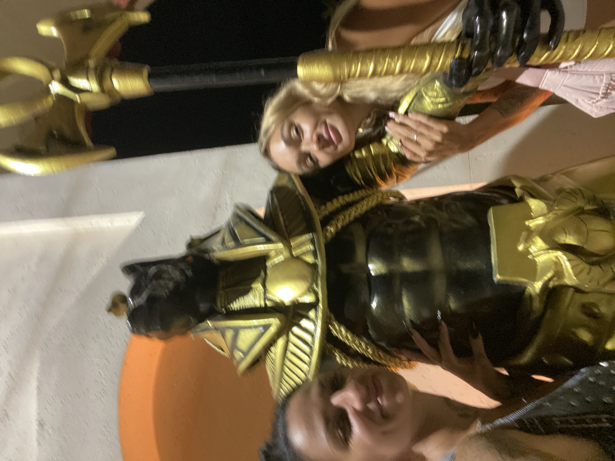 Two women pose with a statue of Anubis at a location in Mũi Né, Vietnam. One woman is blonde, wearing a gold-colored costume, and holding a large prop axe or staff, while the other woman is holding the statue. The statue is mostly black and gold. The background shows a white wall and what appears to be a dark night sky. The woman with blonde hair is sticking out her tongue.