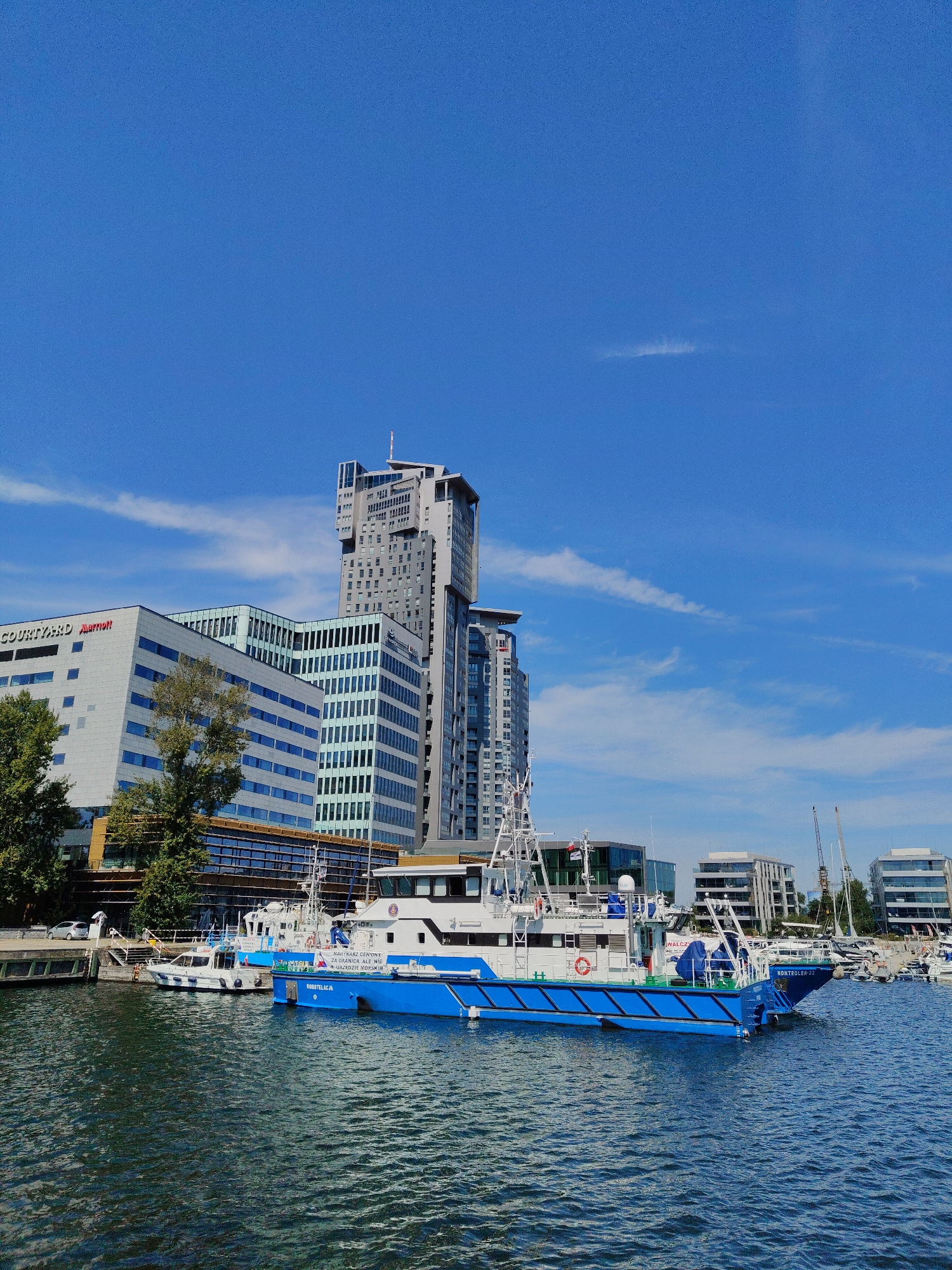 The image depicts the Gdynia waterfront in Poland on a clear, sunny day with a bright blue sky. In the foreground, several boats are docked in the marina. The most prominent is a large blue and white vessel with the name "ROBOSTECJA" and "KONTROLER-22" visible on its hull, suggesting it is a patrol or inspection boat. Another smaller white boat is docked beside it.
In the background, a modern cityscape rises, dominated by the distinctive Sky Tower building, a tall, multi-faceted skyscraper. Adjacent to it are other contemporary office buildings with large windows and geometric designs, including the Courtyard by Marriott hotel. The architecture suggests a developed urban area with a mix of commercial and residential spaces.
Green trees are visible along the waterfront, providing a touch of nature against the urban backdrop. The water in the marina appears calm with gentle ripples, reflecting the blue sky and surrounding structures. The scene conveys a sense of a bustling port city with a modern skyline and active maritime activity.