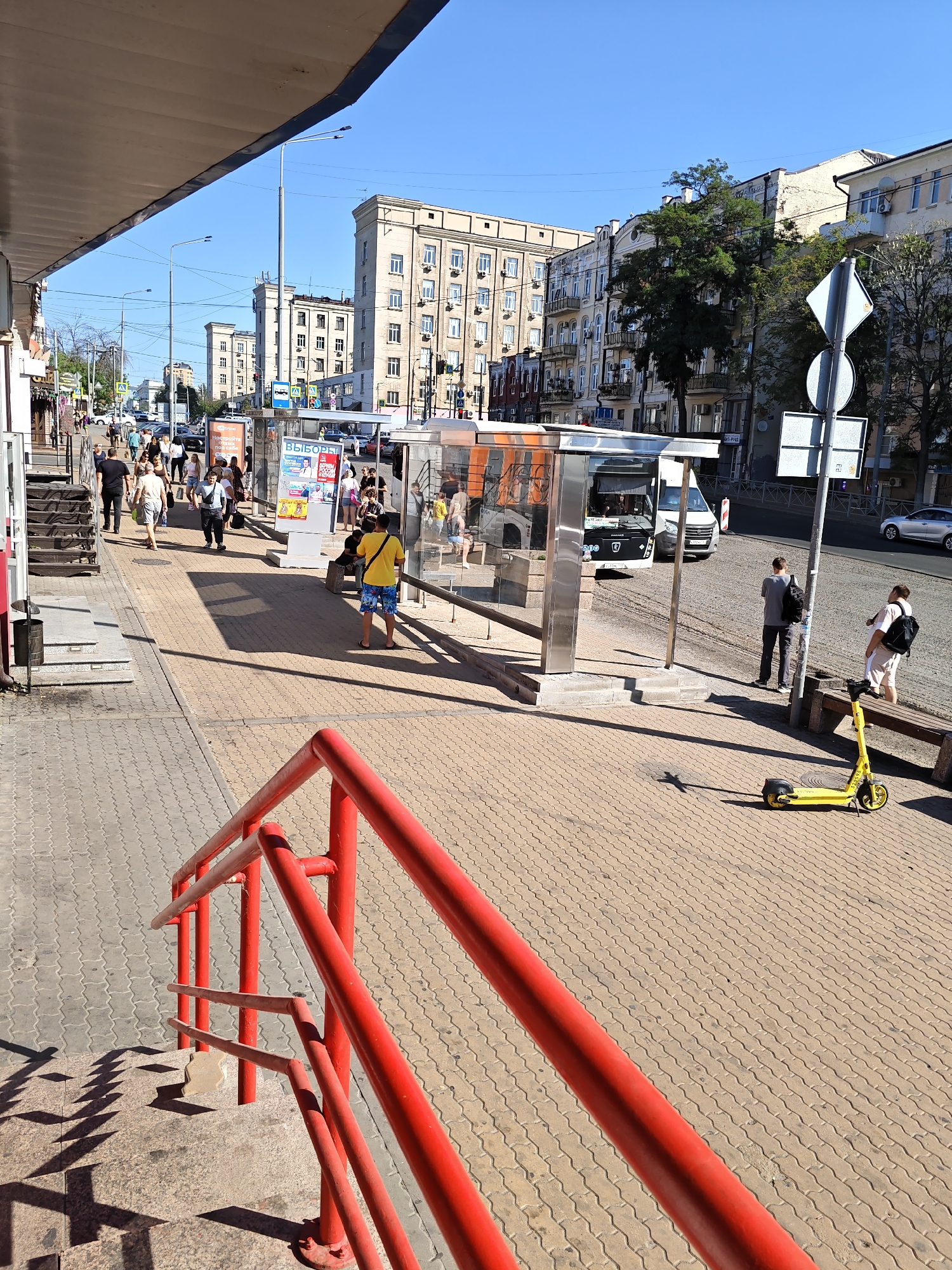 The image shows a street scene in Rostov-on-Don, Russia. A bus stop with a modern glass shelter is visible, with a bus inside and a van parked nearby. Several people are waiting at the bus stop or walking on the sidewalk. A yellow electric scooter is parked on the sidewalk, and a person is sitting on a bench next to it. In the foreground, a red metal railing runs along a set of stairs. The buildings in the background are typical of Soviet-era architecture. The sky is clear and blue, indicating a sunny day. The street is paved with interlocking concrete blocks.