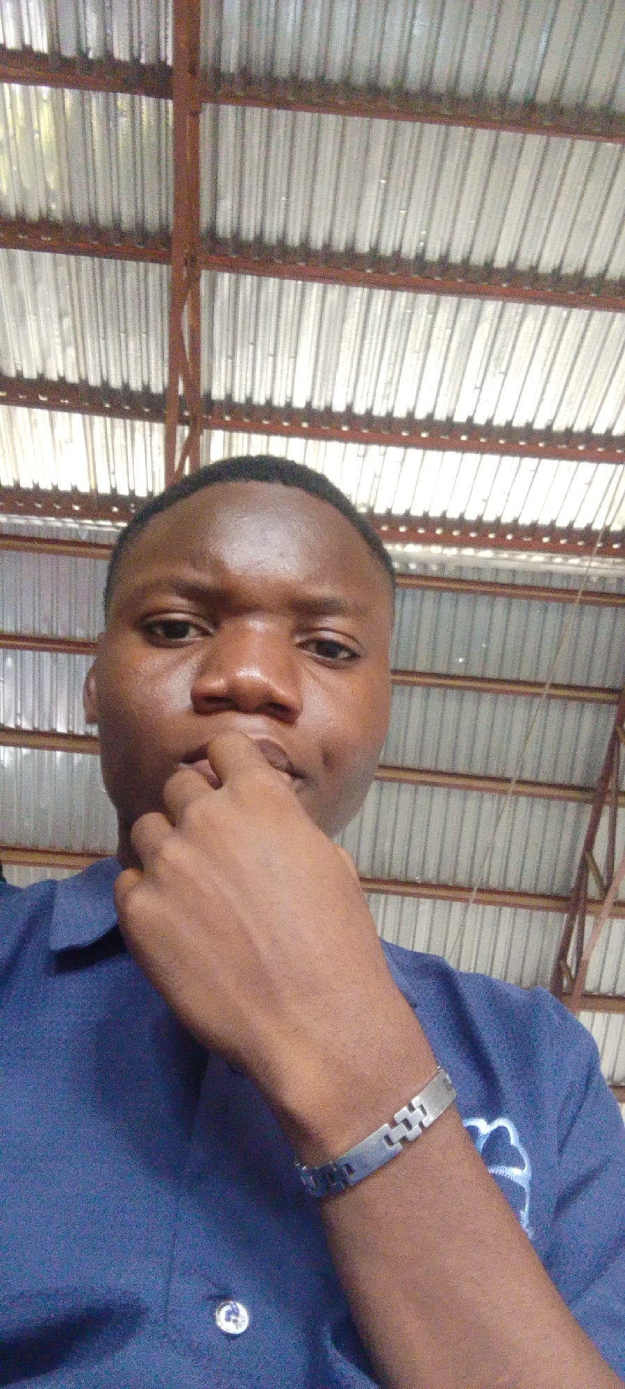 A close-up, waist-up, selfie-style shot shows a young Black person in a blue collared shirt. The person's right hand is raised to their mouth, with their fingers touching their lips. They are wearing a silver-colored link bracelet on their right wrist. The background consists of a corrugated metal roof supported by visible red metal beams, suggesting an indoor or sheltered outdoor industrial or workshop-like environment. The lighting is overhead and appears to be natural, casting a soft glow. The person's face is visible, and they have a thoughtful or contemplative expression.