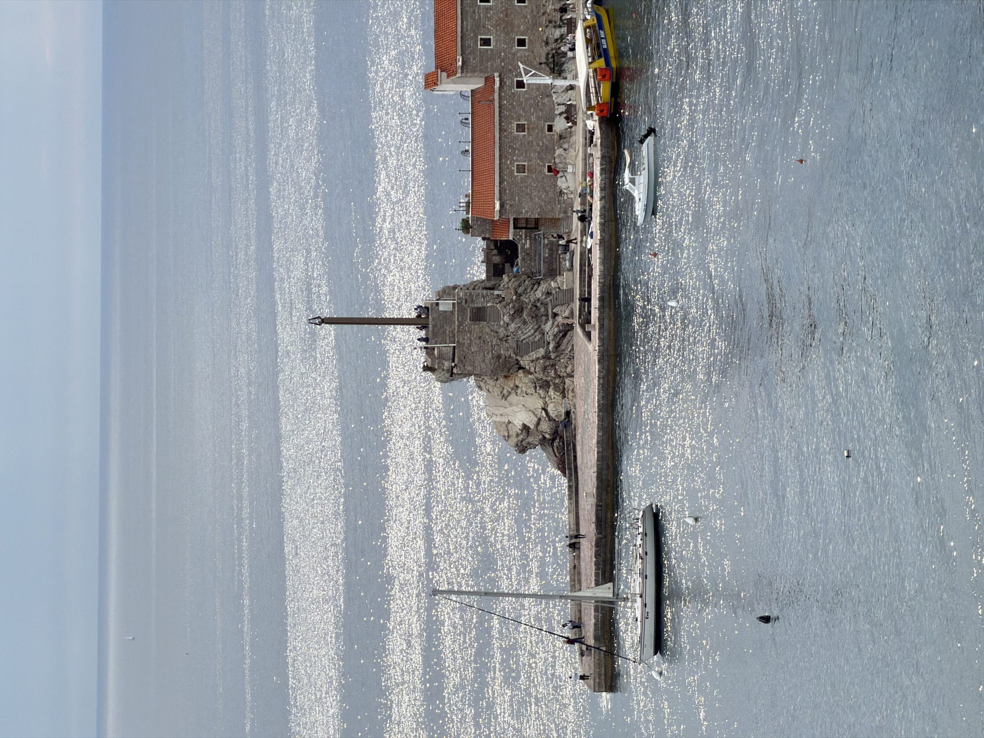 The image depicts a seaside scene in Petrovac na Moru, Montenegro. A stone building complex with a red-tiled roof dominates the center-right. A stone monument stands on a rocky outcrop extending into the sea. People are visible on the monument, the building complex, and along the pier. Several boats, including a sailboat and a tour boat, are docked or anchored near the pier. The sea occupies most of the frame, reflecting sunlight. The sky is visible in the upper left corner.