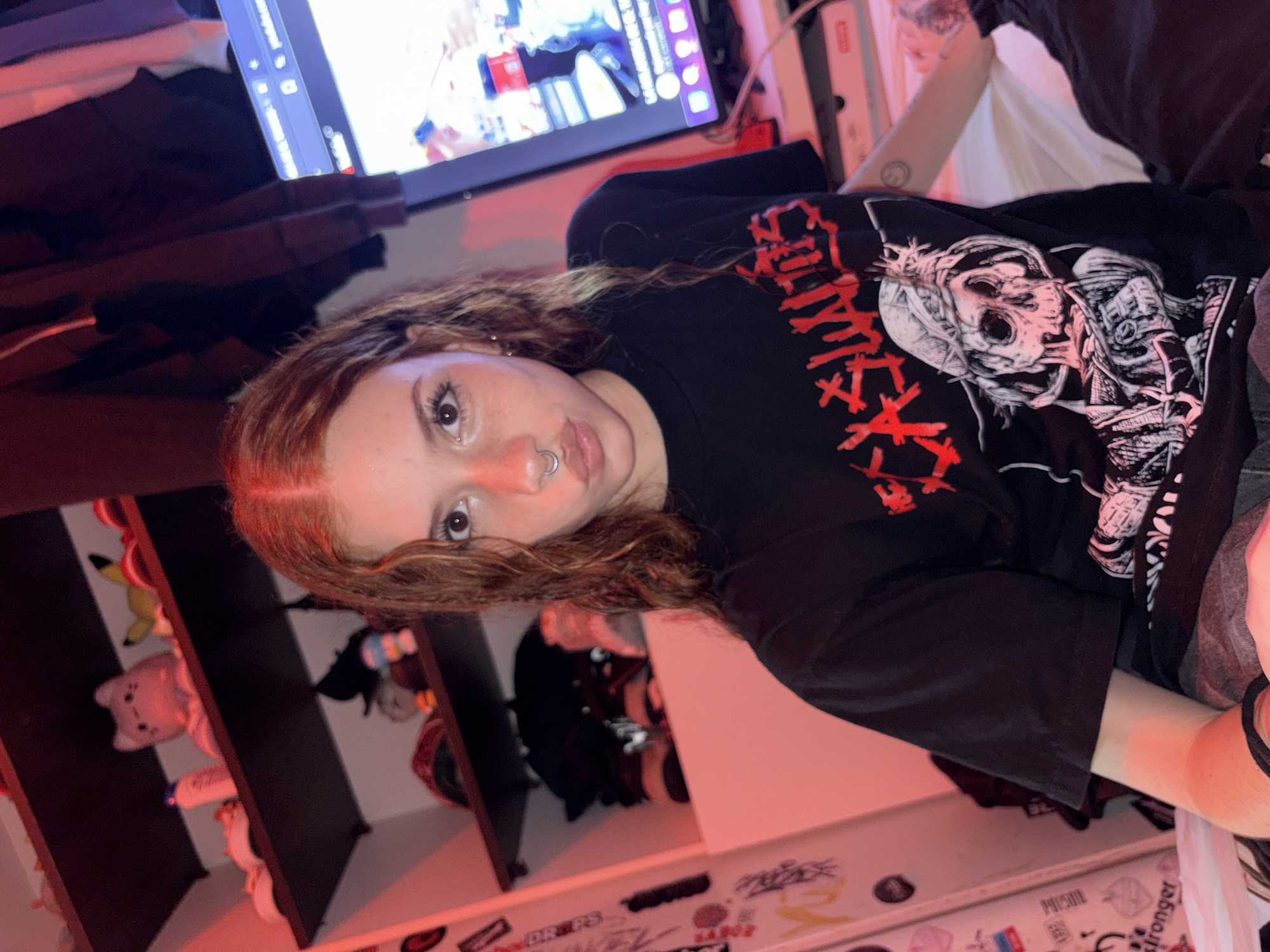A young person, likely female, with reddish-brown, wavy hair, is captured looking directly at the camera with a neutral to slightly pouting expression. She has a silver septum piercing and is wearing a black t-shirt. The t-shirt features an intricate graphic design on the front, depicting multiple skulls and skeletal figures in white and black, alongside stylized red text. Parts of the red text resemble a death metal font, with words like "GORE" and "SKULLS" faintly discernible, suggesting a band logo or genre-specific artwork.
The setting is an indoor space, possibly a bedroom, bathed in a warm, reddish ambient light. In the upper background, a monitor or television screen displays a web browser interface, with the distinct Twitch logo visible in the top left corner, indicating a live stream or video content. To the left, a shelving unit is visible, holding several plush toys, including a yellow Pikachu and a pink Jigglypuff. Below, a white desk or surface is adorned with numerous stickers, some showing partial text like "DR PS" and "STRONGER." The inner arm of another person, revealing a small circular tattoo, is partially visible on the right side of the frame. The image portrays a casual, personal environment, consistent with a room in Buenos Aires, Argentina.