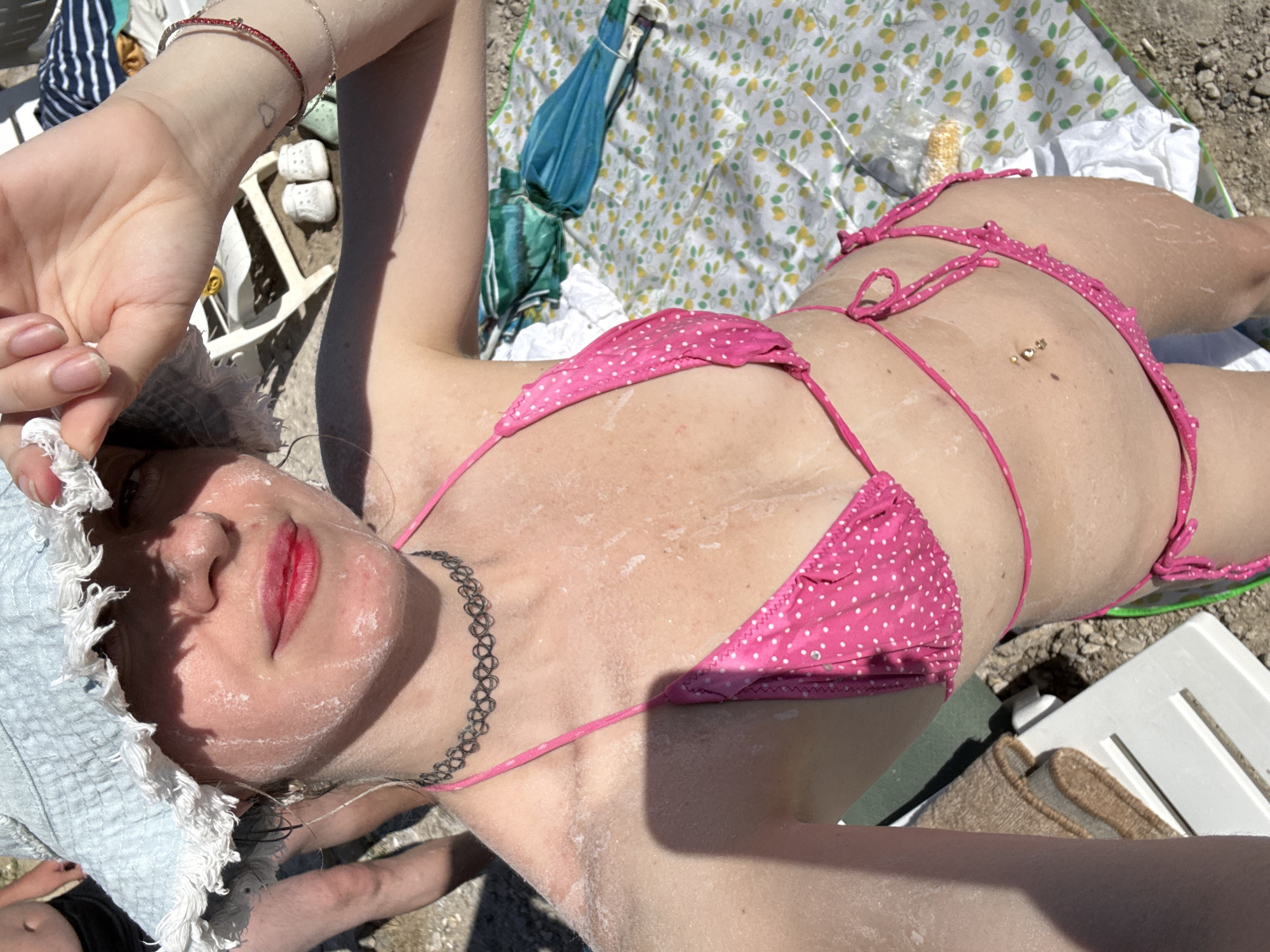 The image depicts a person lying on a beach in Sol-Iletsk, Russia. The subject is wearing a pink polka-dot bikini and a light blue sun hat. Their skin appears to be covered in a white substance, possibly sunscreen or sand. A silver chain necklace is visible around their neck. The person is lying on a patterned blanket, and a beach chair is partially visible. In the background, there are folded umbrellas and some pebbles or sand. The lighting suggests it is a sunny day, likely during daylight hours. The overall scene conveys a sense of relaxation and enjoyment of a summer day at the beach. There is no visible text in the image.