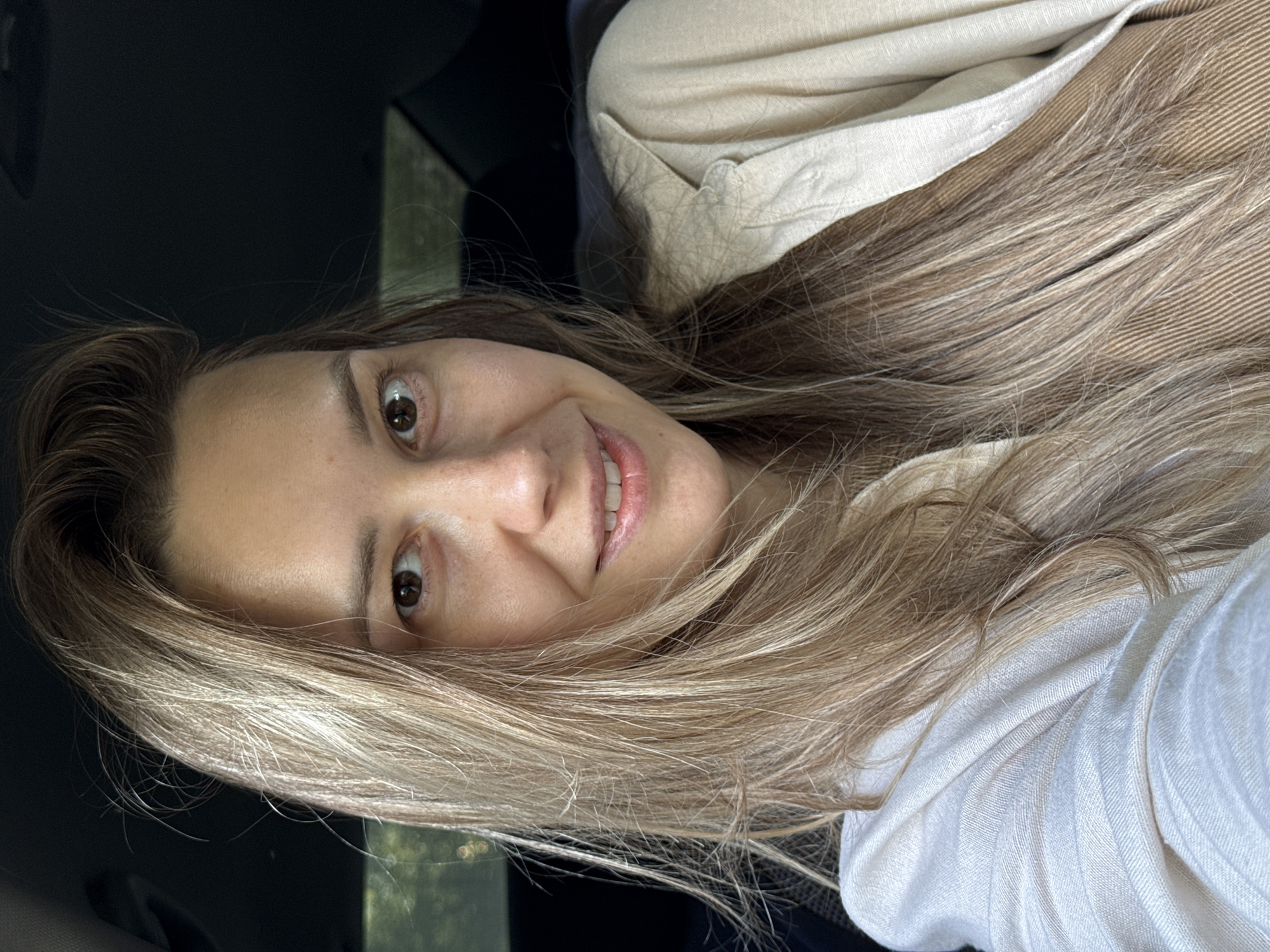 A woman with long, blonde hair smiles into the camera from inside a vehicle. She is wearing a white shirt and a light brown jacket. The environment appears to be a car interior, partially visible through the window. The location in the city of Chaikovsky, Russia is not discernible from the image.