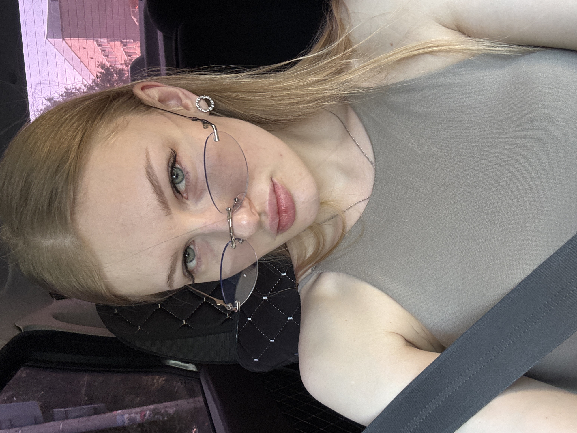 A pale-skinned, blonde woman with blue eyes is in a car in Perm, Russia, as indicated by the urban background visible through the car window. She is wearing a gray top and a seatbelt is across her chest. She is wearing rimless eyeglasses and a small, round earring. The interior of the car has a black quilted pattern on the headrest.