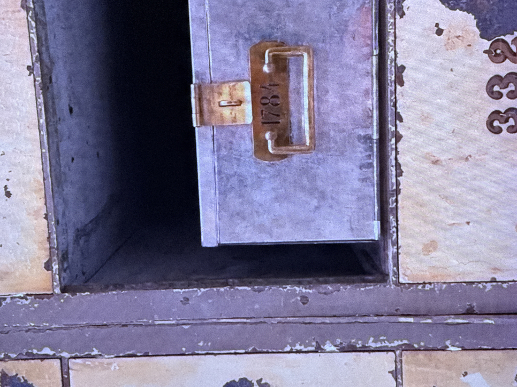 The image shows a close-up view of several old, metallic lockers. One locker is open, revealing a metal box with a latch and a handle. The number "1784" is stamped onto a plate attached to the handle of the box. Another locker, partially visible at the bottom of the image, has the number "332" marked on it. The lockers appear to be made of a weathered, possibly rusted metal, with peeling paint and visible wear and tear. The overall aesthetic suggests an aged and possibly abandoned storage facility. The scene is not identifiable as being in Dubai, United Arab Emirates.