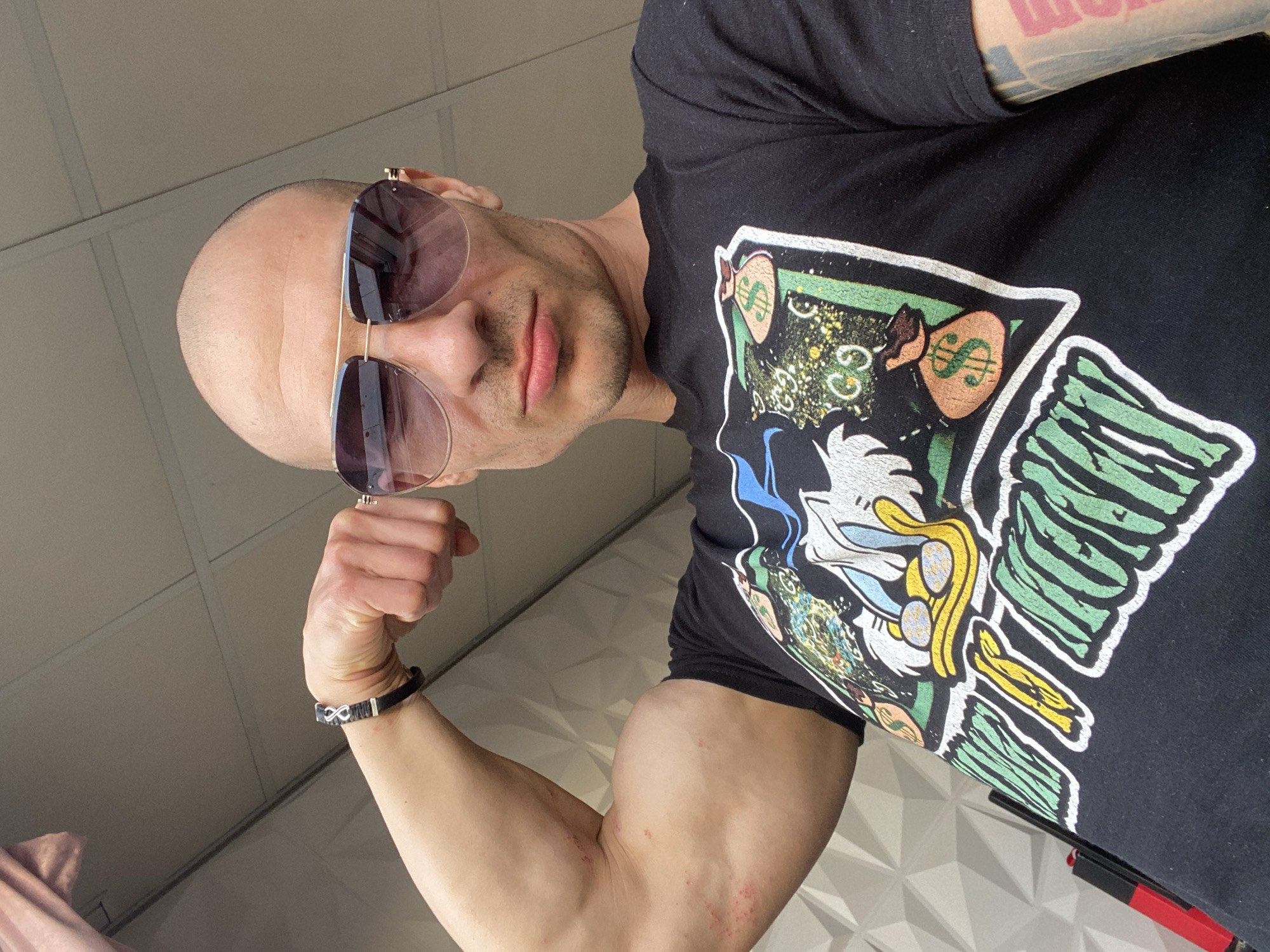 A bald man wearing frameless tinted sunglasses and a black t-shirt with a cartoon money design is flexing his left bicep. He's wearing a black bracelet with a silver infinity symbol. Part of a pinkish fabric is visible in the lower left corner. The background consists of a ceiling and a geometrically patterned wall. There is no specific information to place the subject at the city of Pleven, Bulgaria.