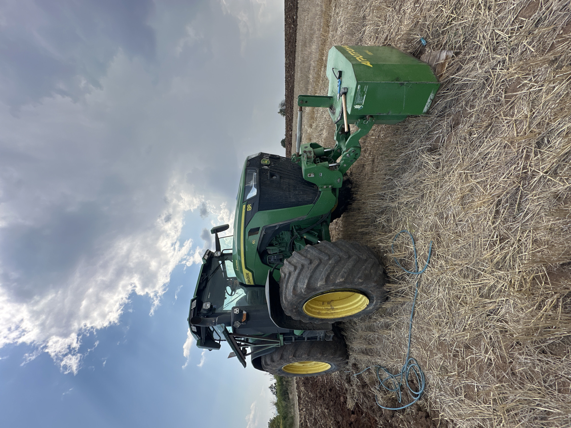 A green John Deere 8R series tractor with yellow wheels is parked in a field of harvested crops. The tractor is equipped with a front-mounted weight box, which has several tools on top, including a hammer and a pair of pliers. A blue cable is coiled on the ground to the left of the tractor. The field is partially plowed, with dark brown soil visible on the left side of the image. The sky is partly cloudy, with dramatic clouds overhead. The location is the city of Kubadin, Bulgaria.
