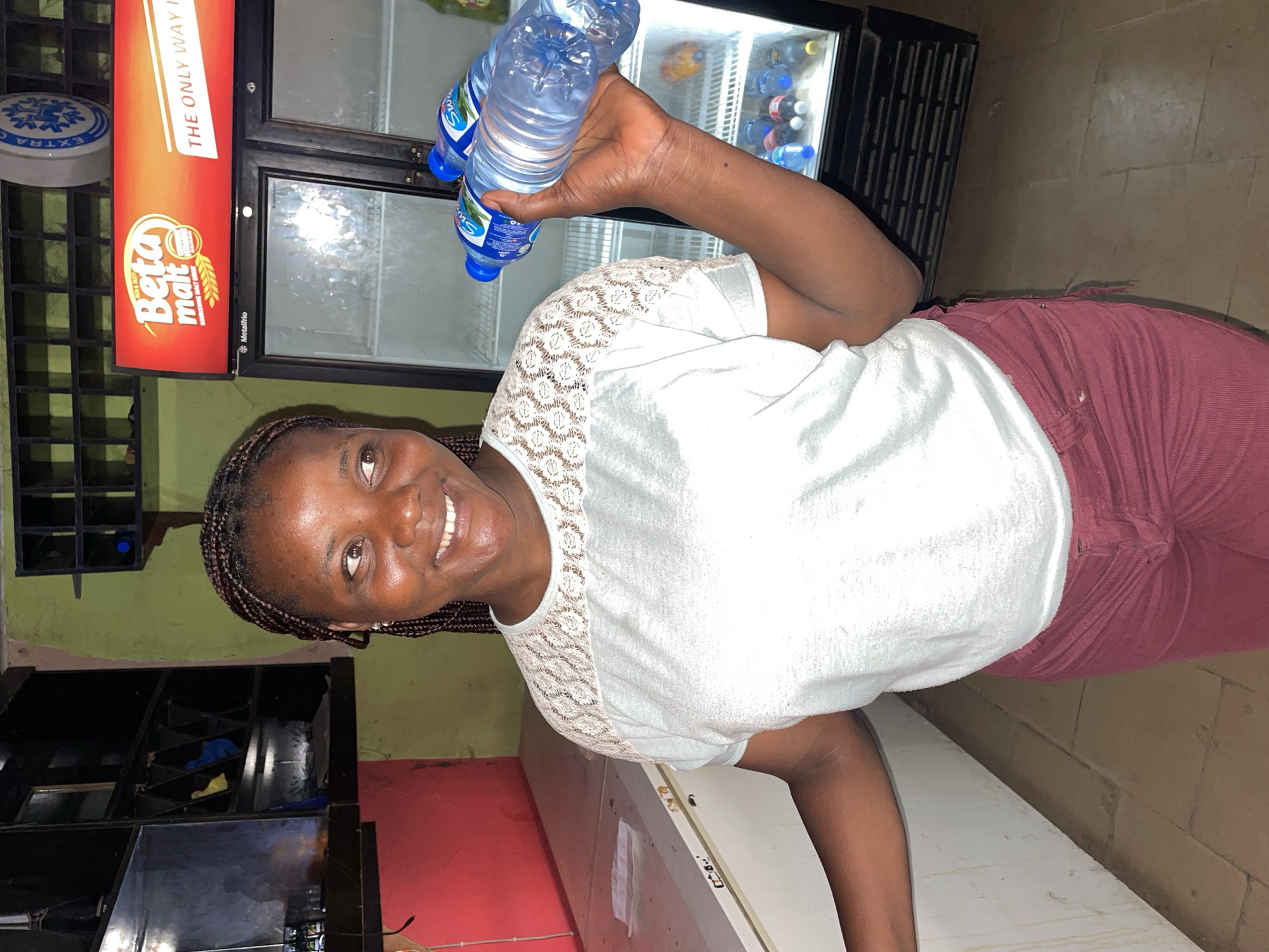 A dark-skinned woman with braided hair is captured smiling broadly at the camera, holding two plastic bottles of water with blue caps and partially visible "Sw" branding (likely Swan) in her right hand. She is wearing a white top with a patterned lace yoke and maroon trousers.
The scene appears to be an indoor commercial establishment, likely a shop or bar, located in Jos, Nigeria. Behind the woman, to her right, is a tall, black commercial display refrigerator, partially stocked with bottled beverages. Adjacent to the fridge is a prominent red-orange advertising panel for "Beta malt," displaying text such as "RICH & TASTY," "NOURISHING MALT DRINK," and a partially visible slogan "THE ONLY WAY T." The "Metalfrio" brand is also visible on the fridge.
On the far left of the image, a green wall is visible, with a black shelving unit or rack mounted on it. A circular sign with "EXTRAC" is also present in the upper left corner. The floor is composed of light brown and beige tiles. The overall lighting suggests an indoor setting, possibly during the evening or in low-light conditions. The woman's cheerful expression is a key emotional element.