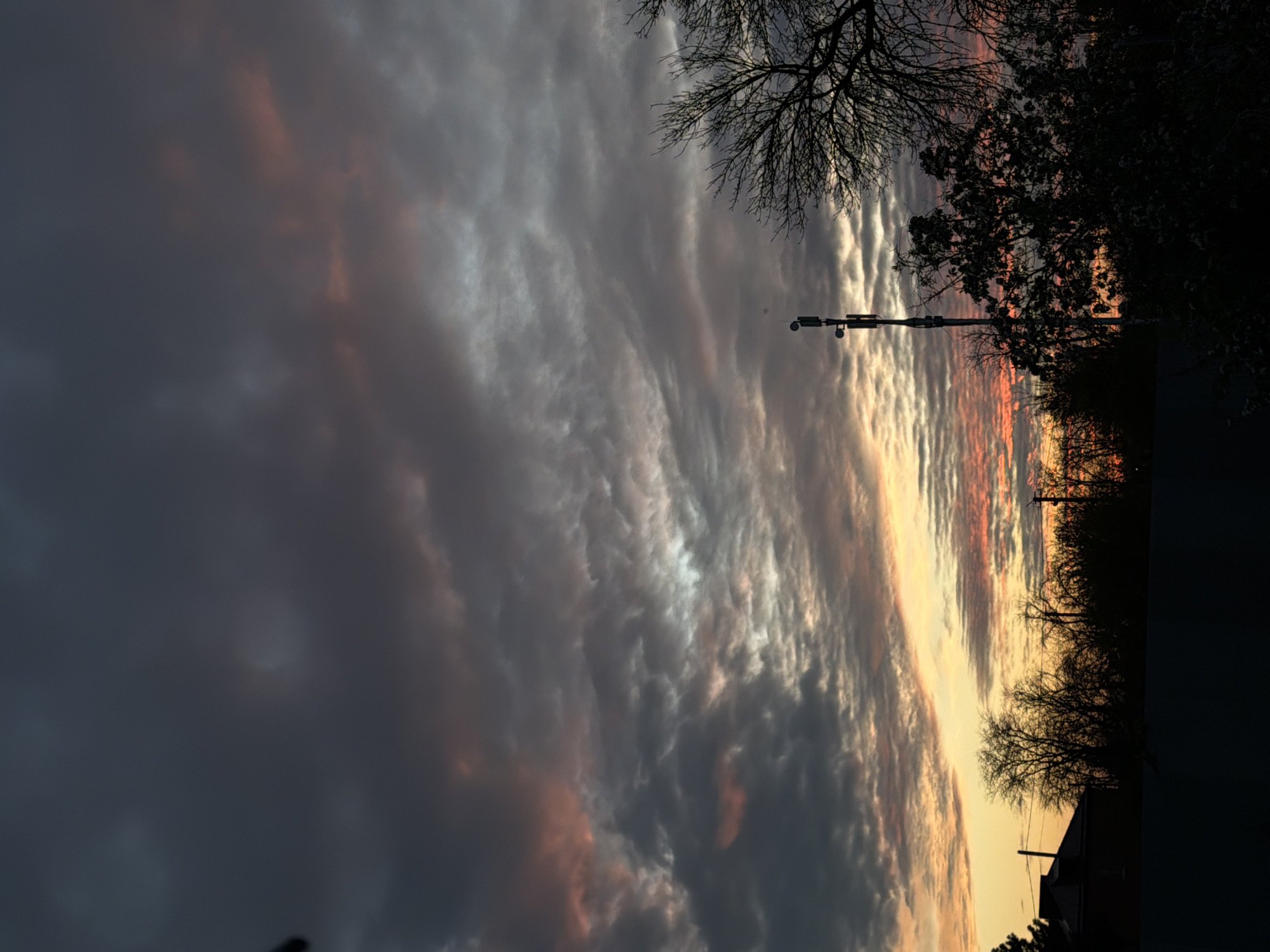 The image depicts a sunset over a cityscape. The sky is filled with striated clouds, transitioning from dark gray on the left to orange and yellow on the right horizon. Silhouetted trees and a utility pole punctuate the skyline. A building is visible in the lower right. The location of the image is not possible to determine from the information provided.