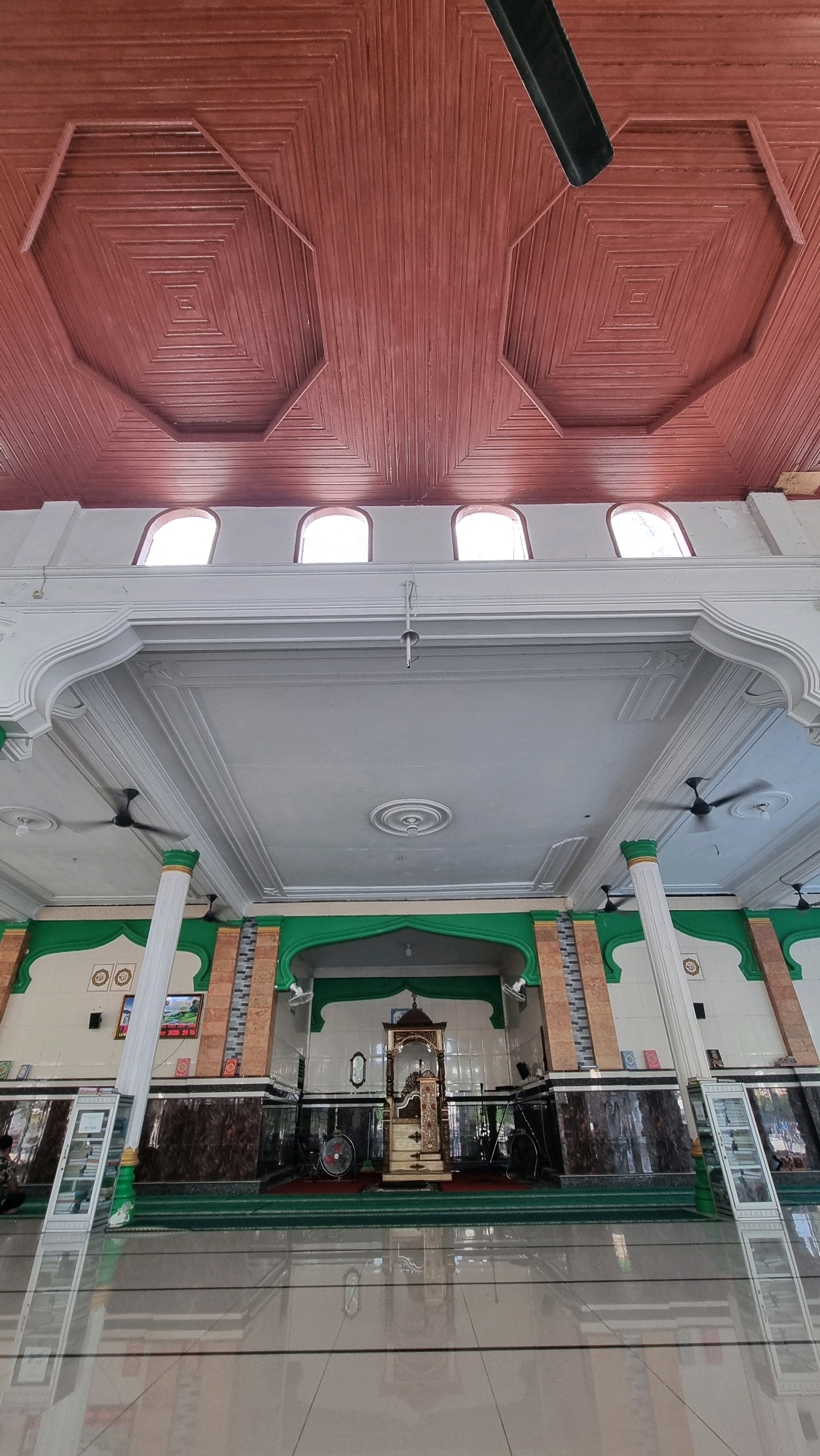 The image shows the interior of a mosque in Blangrambong, Indonesia, focusing on the ceiling and the prayer hall. The ceiling is made of dark red wooden planks arranged in a geometric pattern, with octagonal and square elements. White arched windows are visible at the top of the wall, letting in natural light. Below, the prayer hall features white decorative molding and columns with green accents. The lower section of the wall is adorned with dark marble-like tiles and green arches. In the center of the prayer hall, a wooden pulpit stands on a green carpet. The floor is polished and reflects the interior. Several fans are mounted on the ceiling. The scene is static and appears to be devoid of people actively engaged in prayer or other activities, suggesting it was captured during a quiet period.