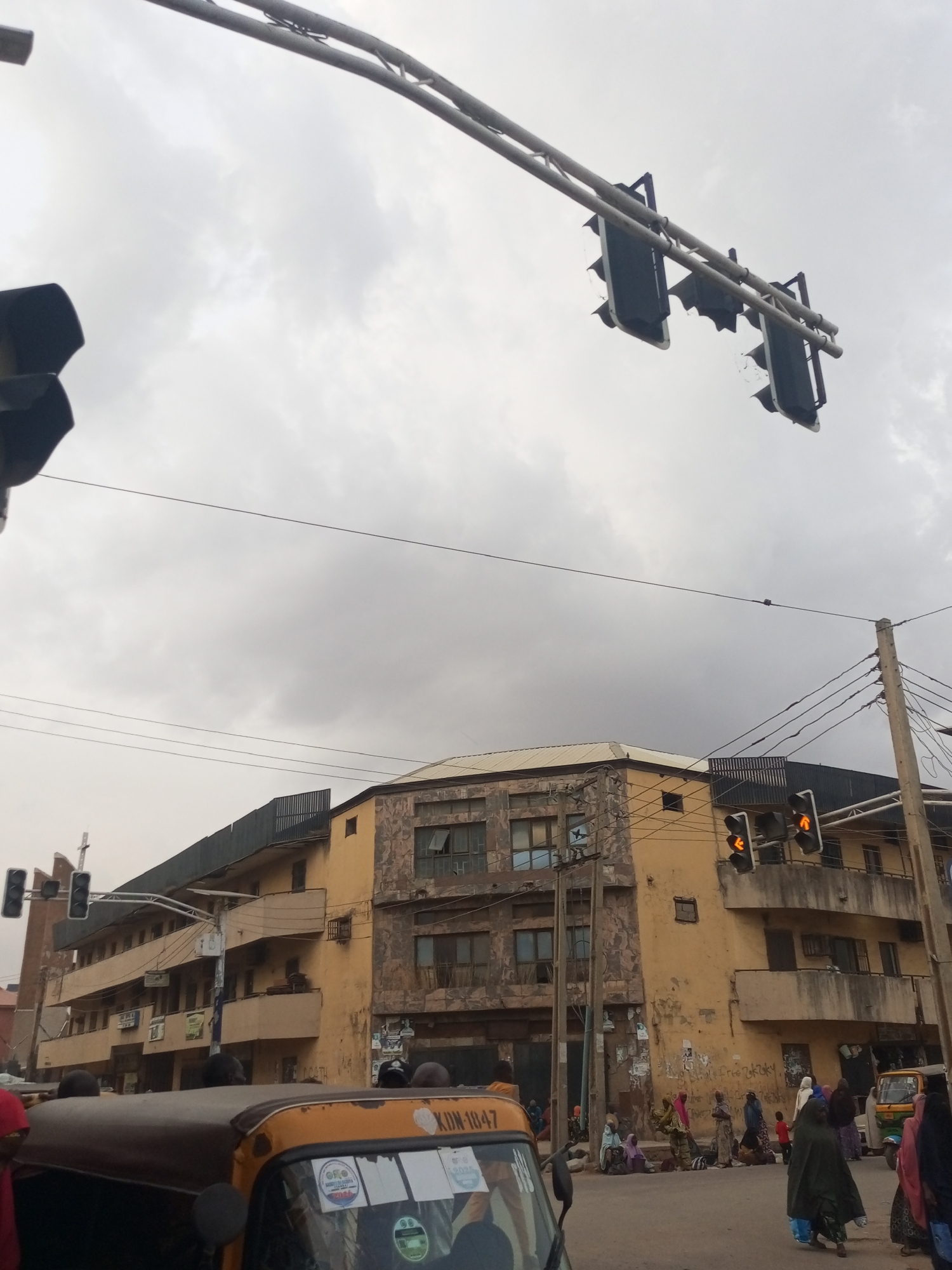 The image depicts a street scene in Maiburiji, Nigeria, under a cloudy sky. Buildings are visible in the background, constructed with multiple floors. The building façades are old and show signs of wear and tear. A metal roof overhangs the tops of the buildings. Traffic lights are suspended above the street on metal bars, with the lights illuminated. A utility pole with multiple wires is situated on the right side of the frame. In the foreground, a yellow auto-rickshaw (a common form of public transport) is partially visible. Groups of people are gathered along the side of the road, some sitting on the ground, others standing. Some are wearing traditional Nigerian clothing.