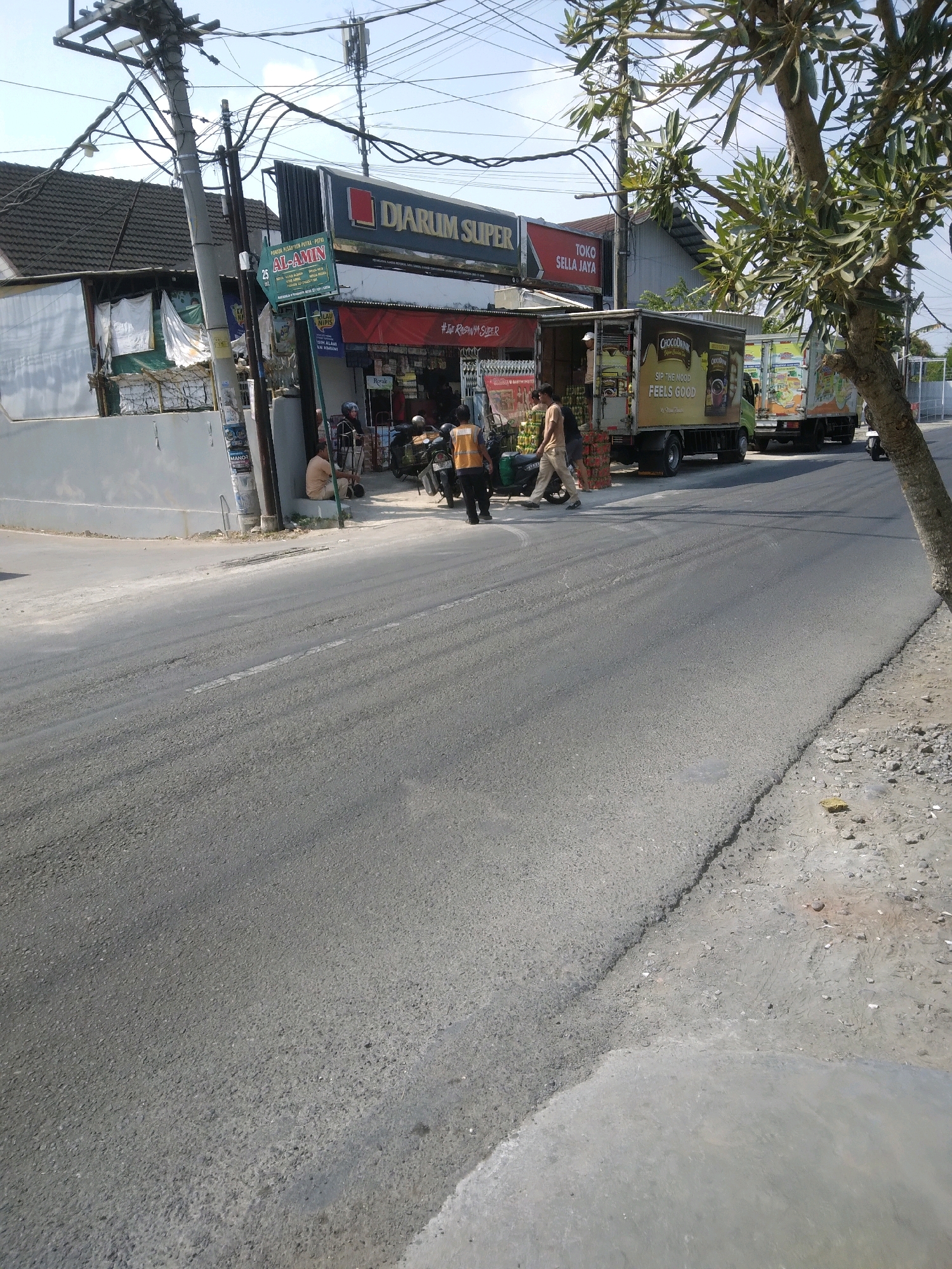 The image shows a street scene in Sokaraja, Indonesia. In the foreground, a paved road is visible with some wear and tear. A small tree with sparse leaves is on the right side of the image. In the background, there is a building with several shops. One shop has a sign that reads "DJARUM SUPER". Another sign above a shop entrance reads "TOKO SELLA JAYA". Two trucks are parked in front of the shops. One truck has a large advertisement on its side featuring the words "CHOCODINK", "SIP THE MOOD", and "FEELS GOOD". Several people are visible in the scene. Two men in orange vests are standing near the trucks. Another man is walking past the trucks. One person is sitting on a low wall in front of a shop. Several motorcycles are parked. The sky is clear and blue.