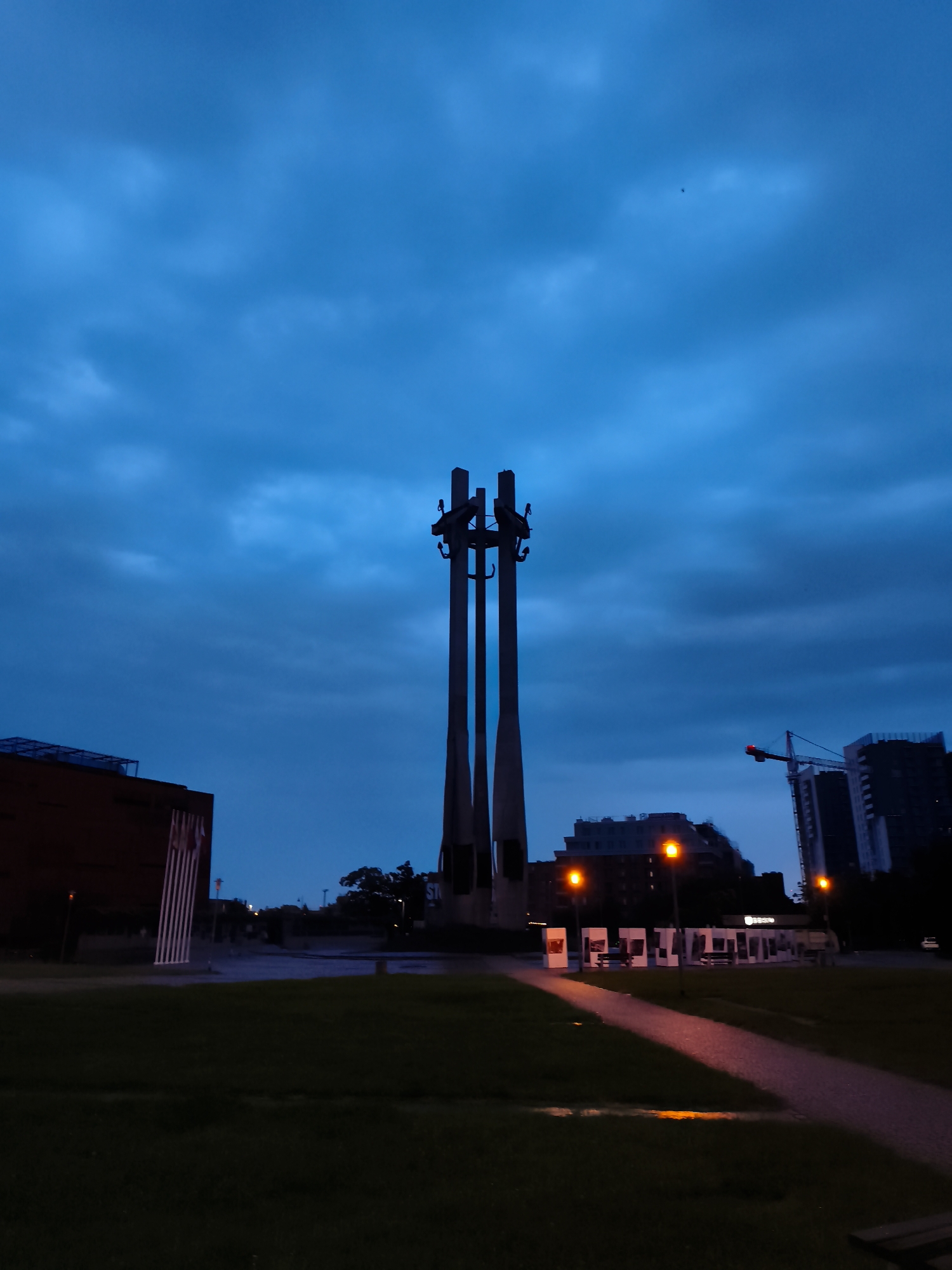 The image depicts the Monument to the Fallen Shipyard Workers of 1970 in Gdańsk, Poland, at dusk. The monument, a prominent symbol of the Solidarity movement, features three large concrete crosses, silhouetted against a deep blue, cloudy sky.
In the foreground, a dark green grassy area stretches towards a paved pathway. The path is illuminated by a few orange streetlights, casting a warm glow and reflecting on what appears to be damp ground. The pathway leads towards a row of white display panels, some of which have images mounted on them, and benches are placed in front of them.
To the left of the monument, a dark, rectangular building with a textured facade is visible. To the right, the cityscape is defined by silhouetted buildings and a construction crane. The overall atmosphere is somber and peaceful, characteristic of the twilight hour in an urban setting. No people are clearly visible in the frame.