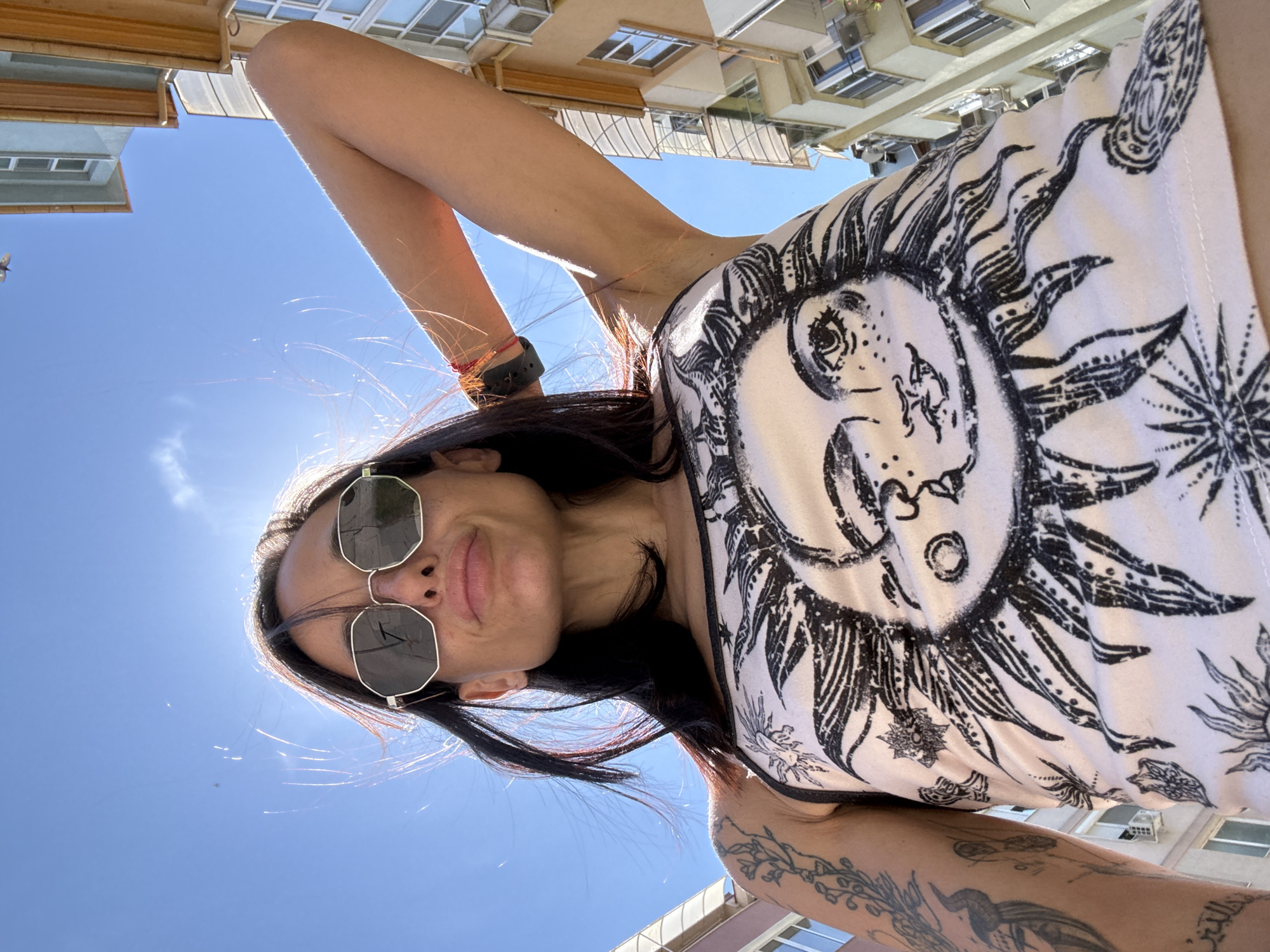 A low-angle, full-body, outdoor, daytime selfie captures a woman posing against a clear blue sky and buildings. The woman, viewed from the chest up, is looking towards the camera, with her head tilted slightly back. She has dark hair, which is partially obscuring her face due to the angle, and is wearing large, hexagonal-shaped sunglasses. Her expression is neutral, with her lips pursed. She is wearing a white tank top adorned with a large black graphic of a sun with a face. The sun's rays extend outwards, and there are smaller celestial motifs around it. The woman's left arm is raised and bent behind her head, showcasing a tattoo on her upper arm. She appears to be in an urban setting, with the sides of buildings visible on the right and left of the frame. The buildings are multi-story structures with windows and balconies. There are no explicit markers indicating the location as Pleven, Bulgaria.