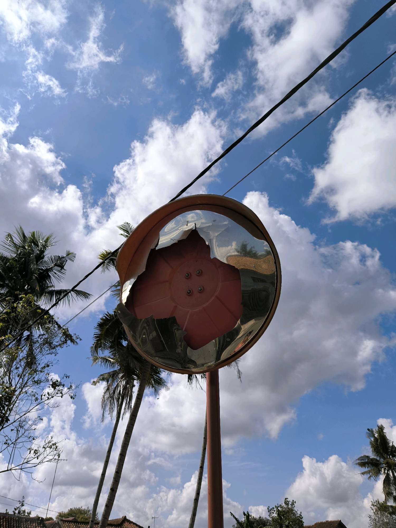 The image captures a wide-angle, low-angle view of a convex traffic mirror with a broken surface, mounted on a reddish-brown pole. The mirror reflects a distorted image of palm trees, buildings with tiled roofs, and a bright blue sky filled with fluffy white clouds. Two dark power lines run horizontally across the upper portion of the frame, passing over the mirror. The scene appears to be outdoors, likely in a tropical or subtropical environment, suggested by the presence of palm trees. The broken state of the mirror indicates potential damage or wear. The backdrop is a vibrant sky, suggesting daylight. The location is specified as Karanganyar, Indonesia. There are no people or animals visible in the frame.