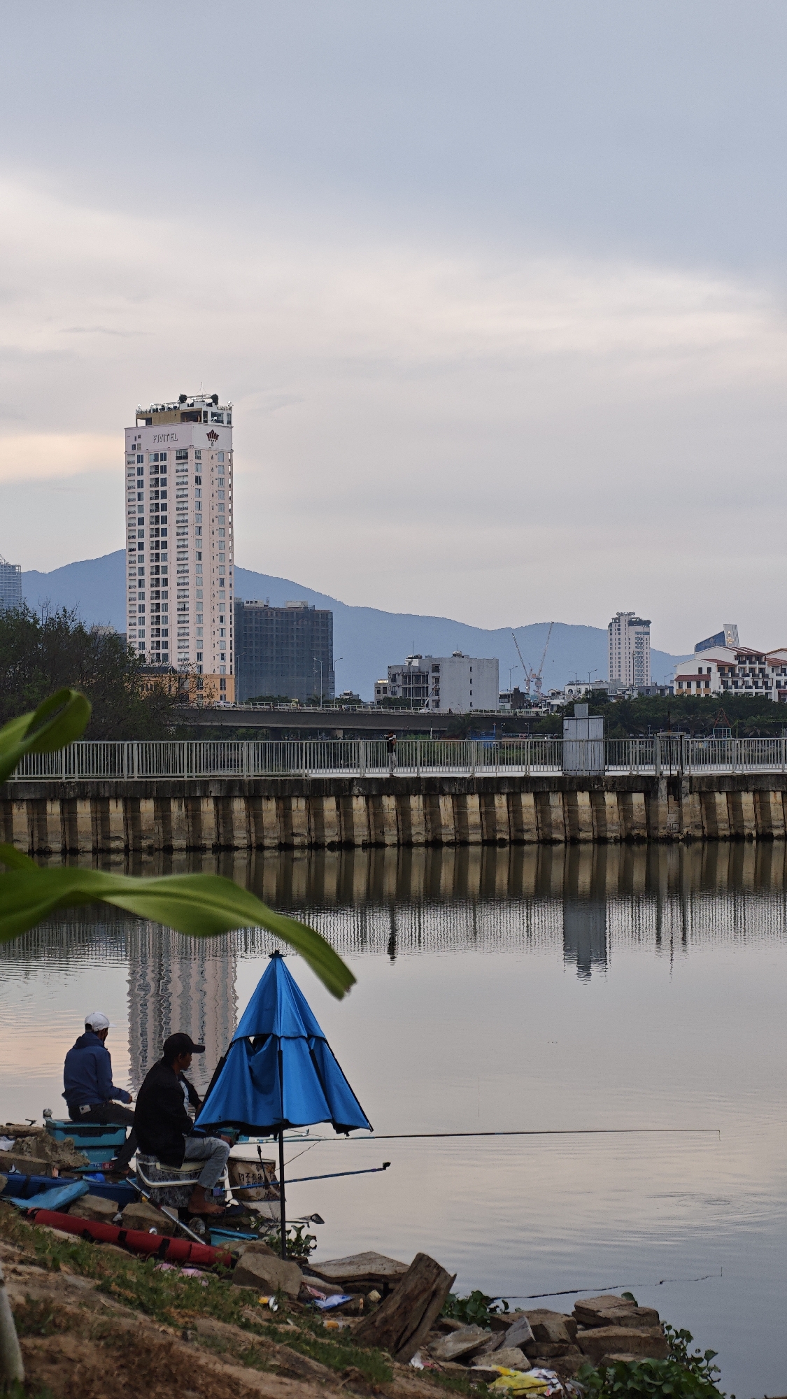 Two men are fishing from the bank of the Han River in Đà Nẵng, Vietnam. One man wears a white cap and blue jacket. The other wears a dark jacket, a dark cap, and is seated on a bucket under a small blue umbrella. Fishing rods are extended over the water. The river bank is littered with trash. In the background, across the river, is a bridge, a tall hotel (identified by its sign as the "Fitel"), and other buildings, with mountains beyond. The sky is overcast. The water reflects the bridge and building.