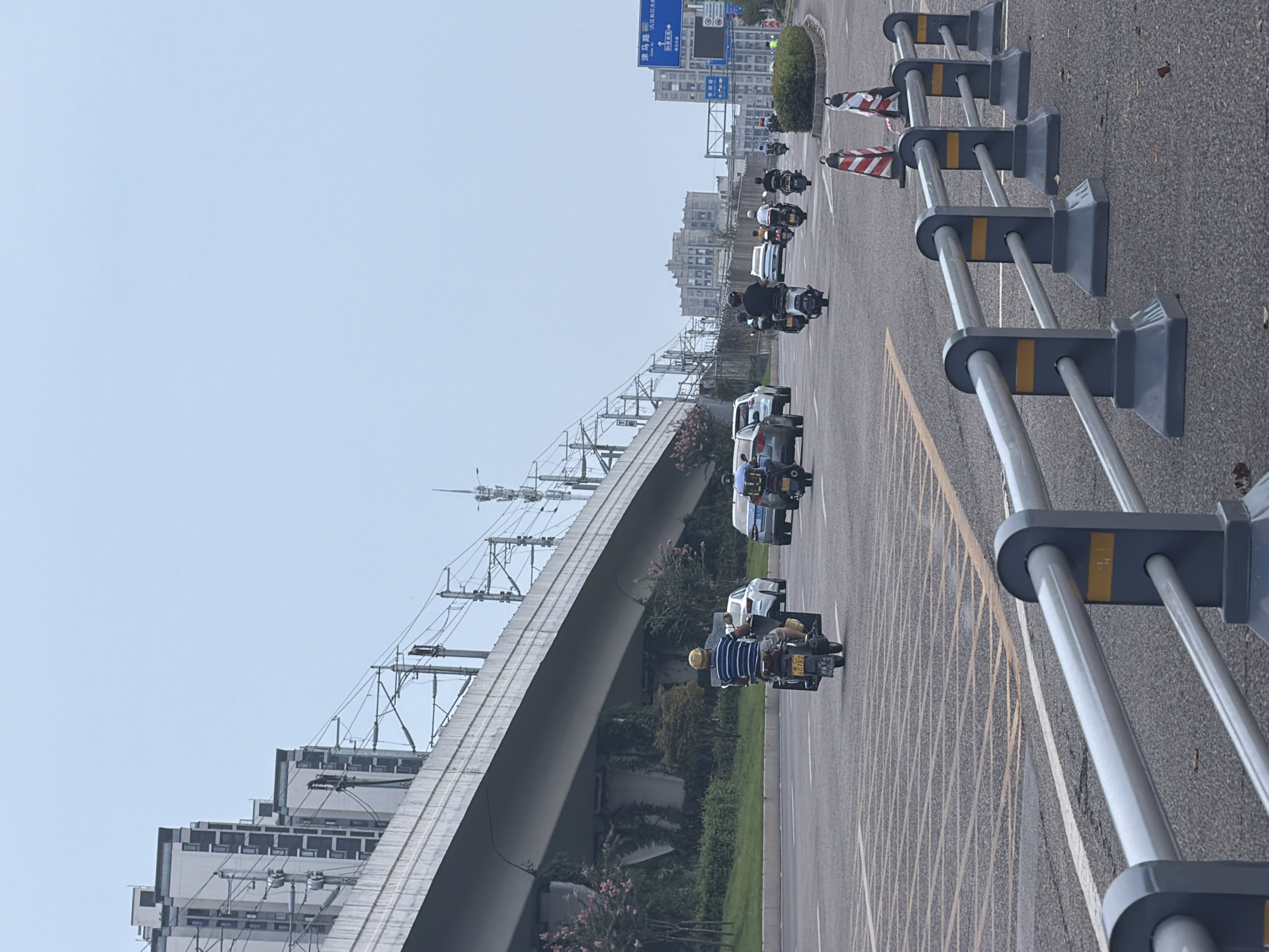 The image depicts a street scene in Jiangjin District, Chongqing, China. A multi-lane road is visible with several motorbikes and cars traveling. In the foreground, a metal barrier with grey posts and silver rails separates the viewer from the road. The road surface is asphalt with yellow and white lane markings, including a section with parallel yellow lines forming a grid pattern. In the background, an elevated structure, likely a highway or train overpass, spans across the scene, supported by concrete pillars. Buildings, including modern apartment complexes, are visible on either side of the road and in the distance. Overhead, power lines and supporting infrastructure for the elevated structure are present. The sky is overcast. A traffic sign with Chinese characters is visible on the right side of the image.