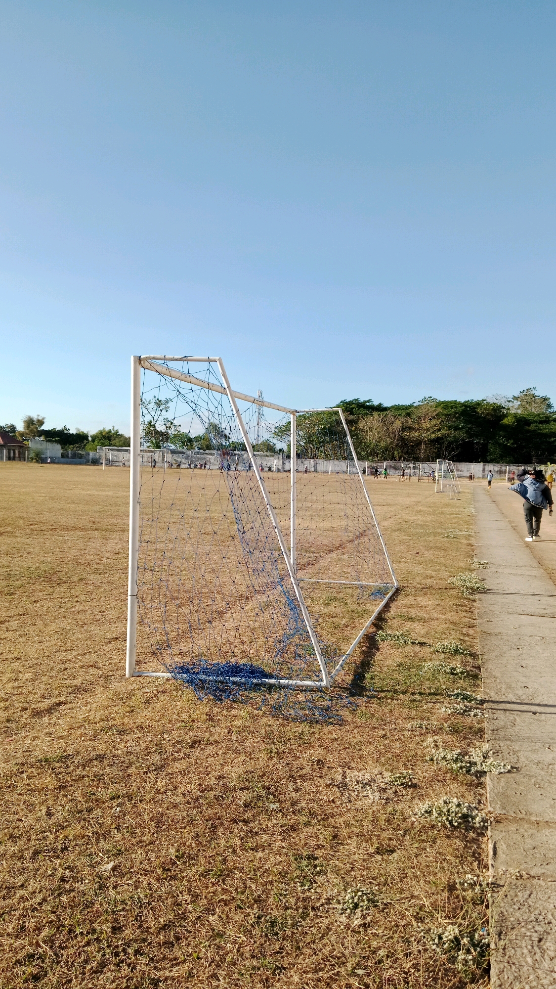 The image depicts a soccer field in Mangkujayan, Indonesia, under a clear blue sky. In the foreground, a white soccer goal with a blue net is positioned on dry, brown grass. The net is partially collapsed. A concrete path runs along the right side of the field, bordered by sparse green vegetation. In the mid-ground and background, other soccer goals are visible, and several individuals, including a person walking away from the camera on the path, are present on the field, some appearing to be engaged in activity. Trees and a fence line the perimeter of the field. The overall impression is a somewhat parched, outdoor sports environment.