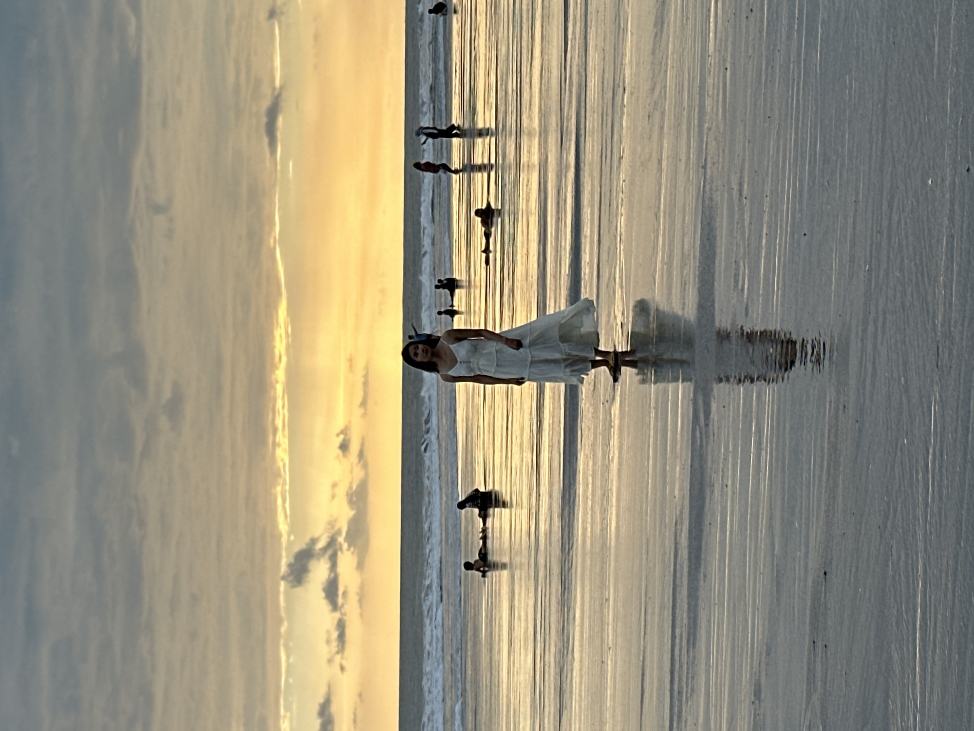 A young woman in a flowing white dress stands on a wet beach at sunset in North Kuta, Indonesia, her reflection clearly visible on the water's surface. The sky above is a gradient of soft grays and warm yellows, with scattered clouds. The ocean waves are breaking gently in the background, and other people, silhouetted against the light, are scattered across the beach and in the shallow water, some sitting, some standing, and some wading. The overall scene evokes a serene and peaceful atmosphere.