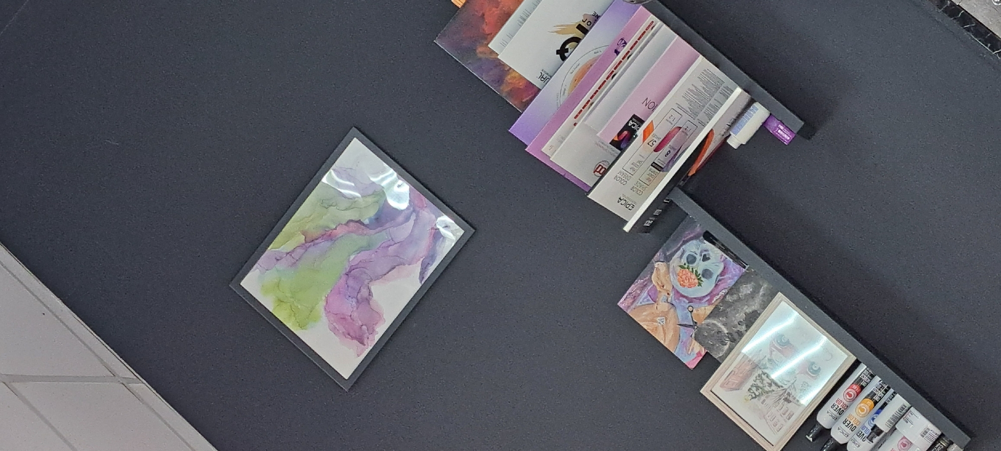 The image depicts a wall with several framed artworks and a shelf with art supplies and books. In the upper left is a small framed artwork with abstract green and purple watercolor splashes. Below and to the left of this is a shelf displaying a small framed picture of what appears to be a vintage car in a snowy scene, and several tubes of art markers. To the right of this shelf, and slightly higher, is another shelf angled upwards, holding several books or art swatch books with colorful covers, and a small bottle with a purple cap. To the left of the angled shelf is a small framed artwork featuring a skull adorned with a flower and a person reclining, with scissors nearby. The wall is dark gray. The image does not provide any information about its location in Yessentuki, Russia.