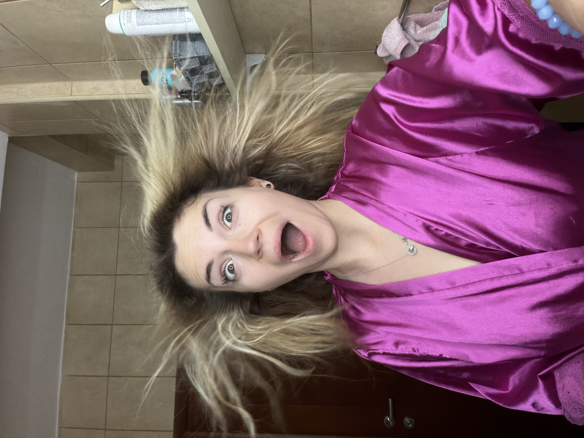 This image is a medium close-up portrait of a young woman, rotated 90 degrees counter-clockwise. The woman, who has light skin, wide brown eyes, and light brown/blonde hair, displays an exaggerated expression of surprise or playfulness with her mouth open in an "O" shape. Her hair appears fanned out and standing on end around her head, creating a dynamic effect. She is wearing a shiny, vibrant magenta or fuchsia satin robe, and a delicate silver necklace with a small pendant. A small black stud earring is visible in her left ear.
The setting appears to be an indoor room, likely a bathroom or dressing area, indicated by the light brown tiled floor or wall and various toiletries. Behind her head (to the left in the rotated image), a shelf holds a white bottle (partially visible text includes "shampoo" and "conditioner," likely a Dove product), a smaller blue/green bottle, and a grey and white checkered towel or fabric. To the right of her body, a dark brown door with a metallic handle is visible, and on an adjacent surface, a light pink towel and a blue beaded accessory are present. The overall atmosphere conveyed by the woman's expression is one of lightheartedness and amusement. The location context provided is Budva, Montenegro.
