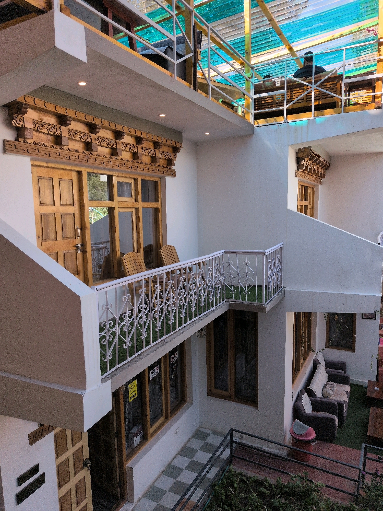 The image depicts the interior courtyard of a multi-story building, likely a guesthouse or hostel, in Leh, India. The architecture features traditional Ladakhi wooden carvings above the windows and doors. The courtyard is open to the sky, with a translucent, colored roof structure visible in the upper portion of the image.
On the first floor, a wooden door and a window with multiple panes are prominent. Two wooden chairs are placed on a small balcony in front of this window, which is decorated with a white metal railing and artificial grass. The door appears to be made of several panels and has a lock.
The second floor features a walkway with a similar white metal railing, overlooking the first floor. On this level, two individuals are seated on a wooden bench, partially obscured by the railing and the roof structure. The roof is supported by wooden beams and covered with corrugated, colored plastic sheeting, allowing natural light to filter through.
Further into the courtyard, on a lower level accessible by stairs, there is an outdoor seating area with dark sofas, a coffee table, and what appears to be a red plastic dustbin. A checkered tile floor is visible in this area. The walls are painted white. Signs indicating "No Smoking" are visible on some windows. The overall impression is a blend of traditional local aesthetics and functional, modern amenities.