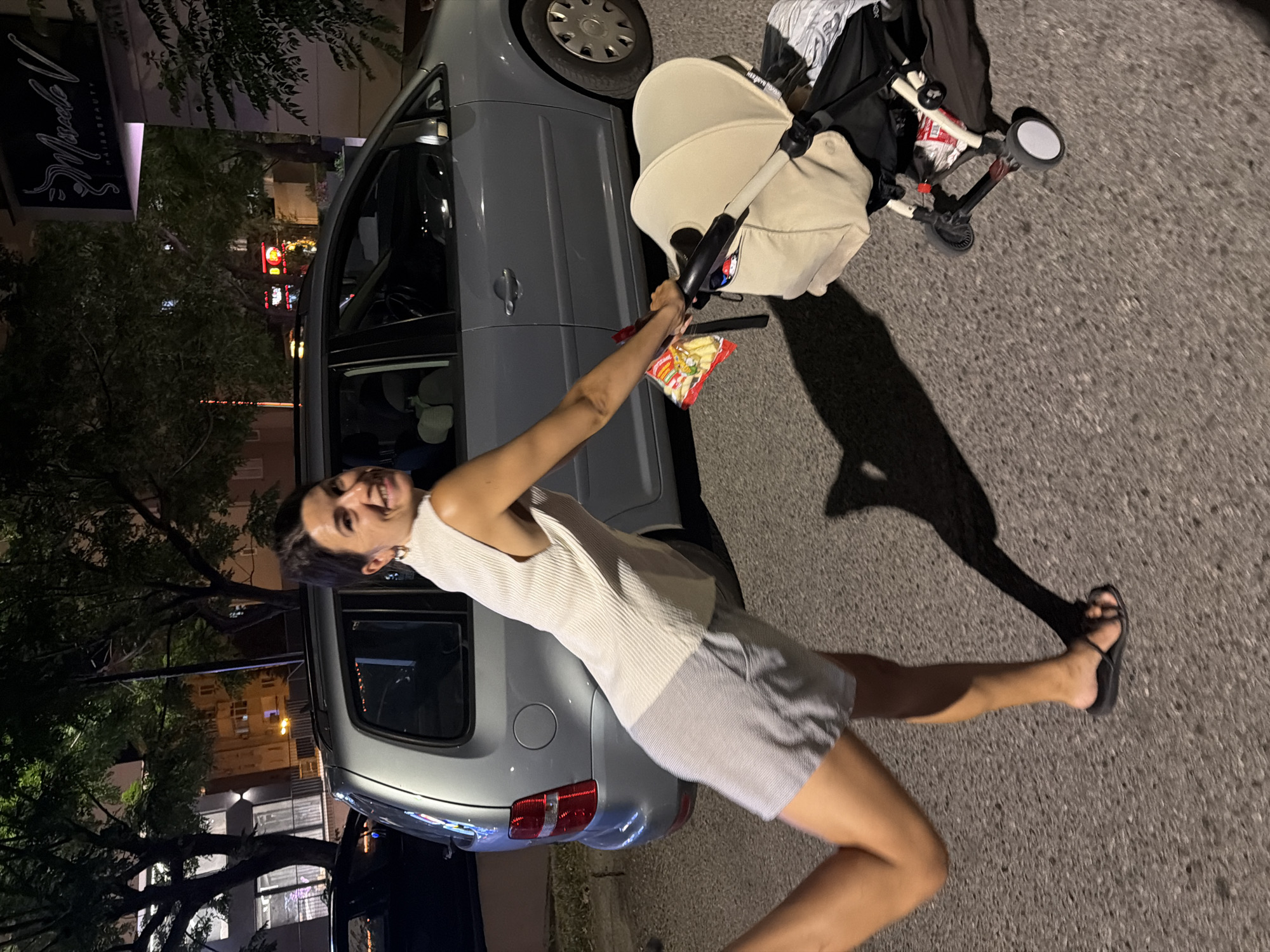 A woman with dark hair is leaning out of a car door, holding a bag of chips and smiling. She is wearing a white sleeveless top and grey shorts. A stroller is parked next to the car. The scene appears to be on a street at night, with streetlights and building lights visible in the background. Trees are also present, casting shadows. The woman's expression suggests happiness or amusement.
