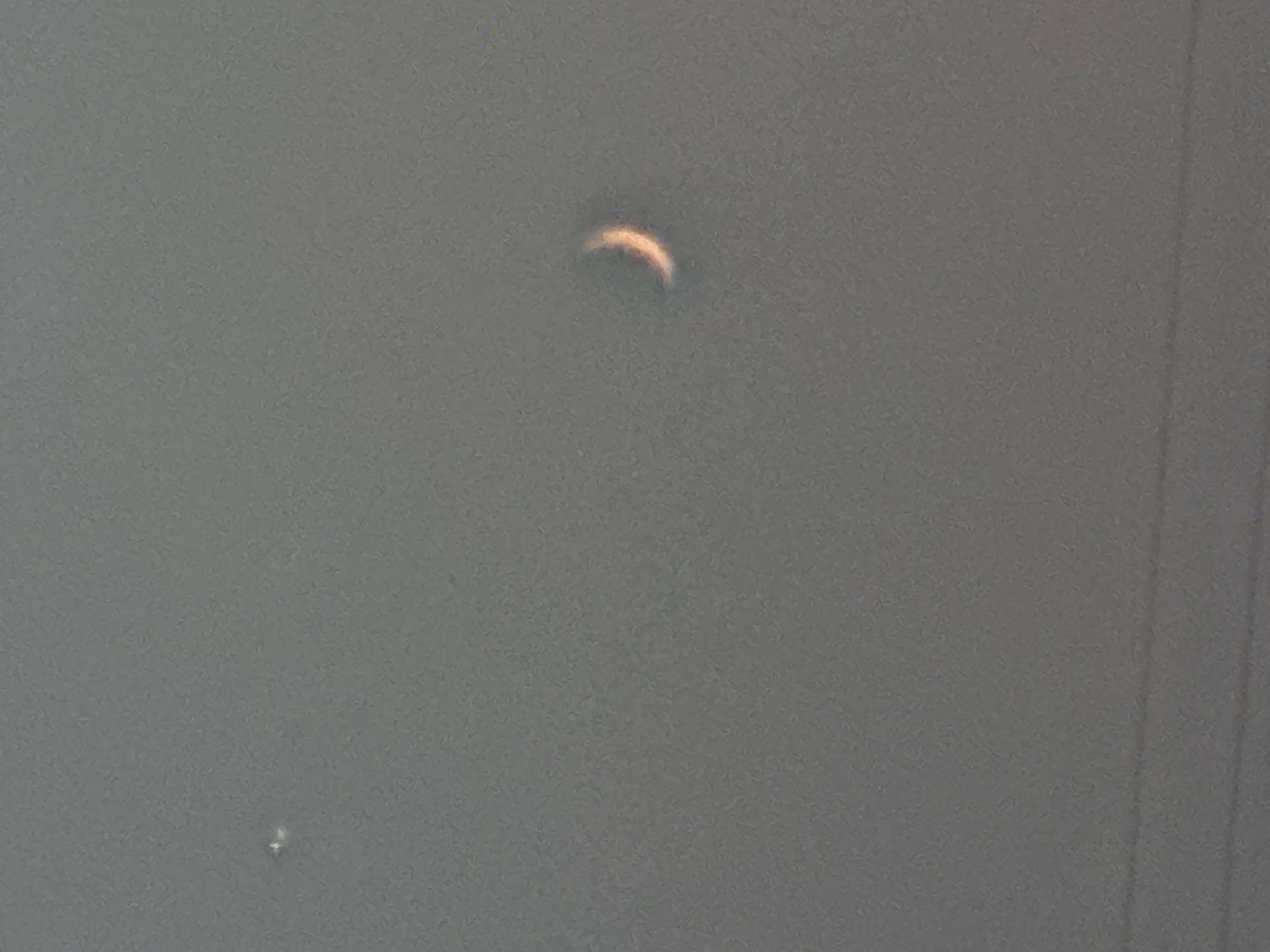 The image shows a hazy, grey sky with two celestial objects. On the left, there is a small, bright white object that appears to be a star or planet. On the right, there is a crescent-shaped object, possibly the moon or a solar eclipse, illuminated with a warm, orange glow. The bottom of the image shows faint horizontal lines, which could be telephone wires or power lines. There are no people, buildings, or other discernible elements of the city of Biysk, Russia visible in this image.