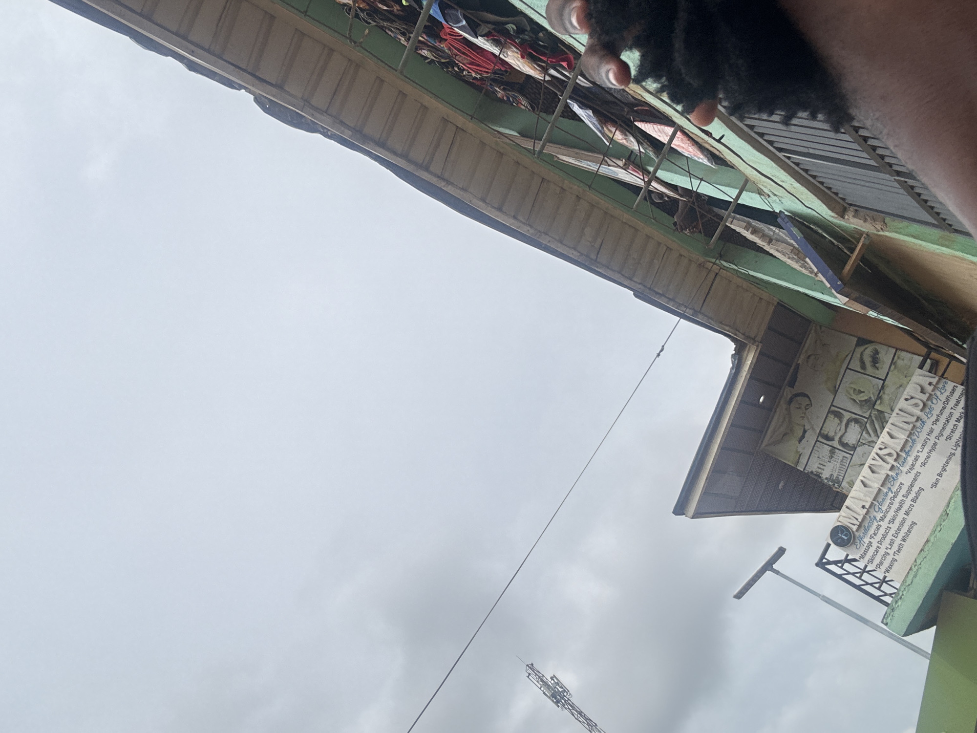 The image, taken at an angle, shows a building with a sign reading "MAKKYSKIN SPA" in Ihiagwa, Nigeria. The sign advertises various services, including skincare, massages, manicures/pedicures, and hair treatments. A cell tower and power lines are visible in the background against an overcast sky. Clothing and other items are haphazardly stored above the building's roofline. The photograph appears to be taken from a low angle, looking upwards toward the building and the sky. A portion of a person's arm and possibly hair are visible in the upper right corner of the frame.