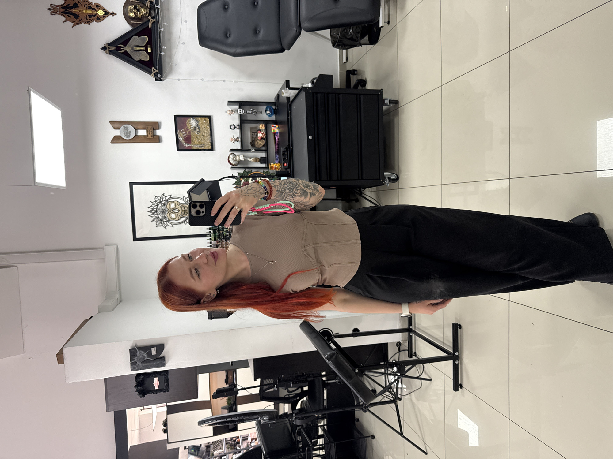 A woman with red hair stands in a tattoo studio. She is wearing a beige top and black pants, and she is holding a smartphone in her left hand, taking a selfie. Her right arm is tattooed. The tattoo studio has white walls and a tiled floor. There is a tattoo chair, a black tool chest, and various framed artworks on the walls. Another tattoo station is visible in the background. The location is likely in Rostov-on-Don, Russia.
